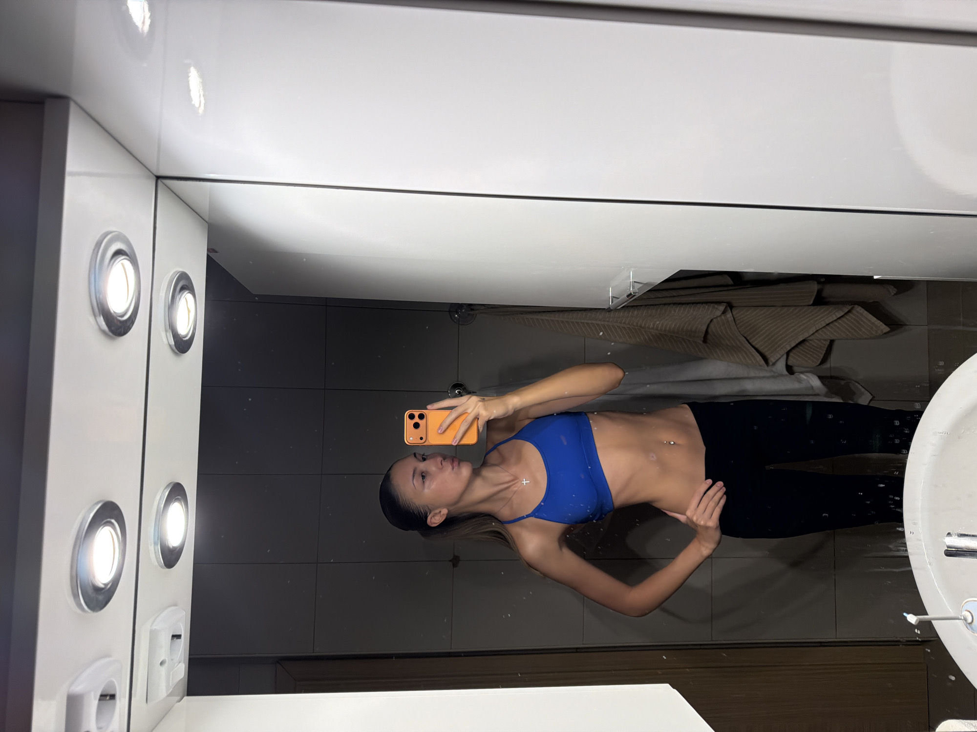 A young woman stands in front of a mirror, taking a selfie. She is wearing a blue sports bra and black leggings. She has a slim build and a silver cross necklace with a thin chain. Her hair is pulled back in a ponytail. She is holding an orange smartphone in her right hand. The background appears to be a bathroom with tiled walls and a white vanity. There are several recessed ceiling lights above the mirror, and a towel hangs on a rack to the right. The overall lighting is dim.