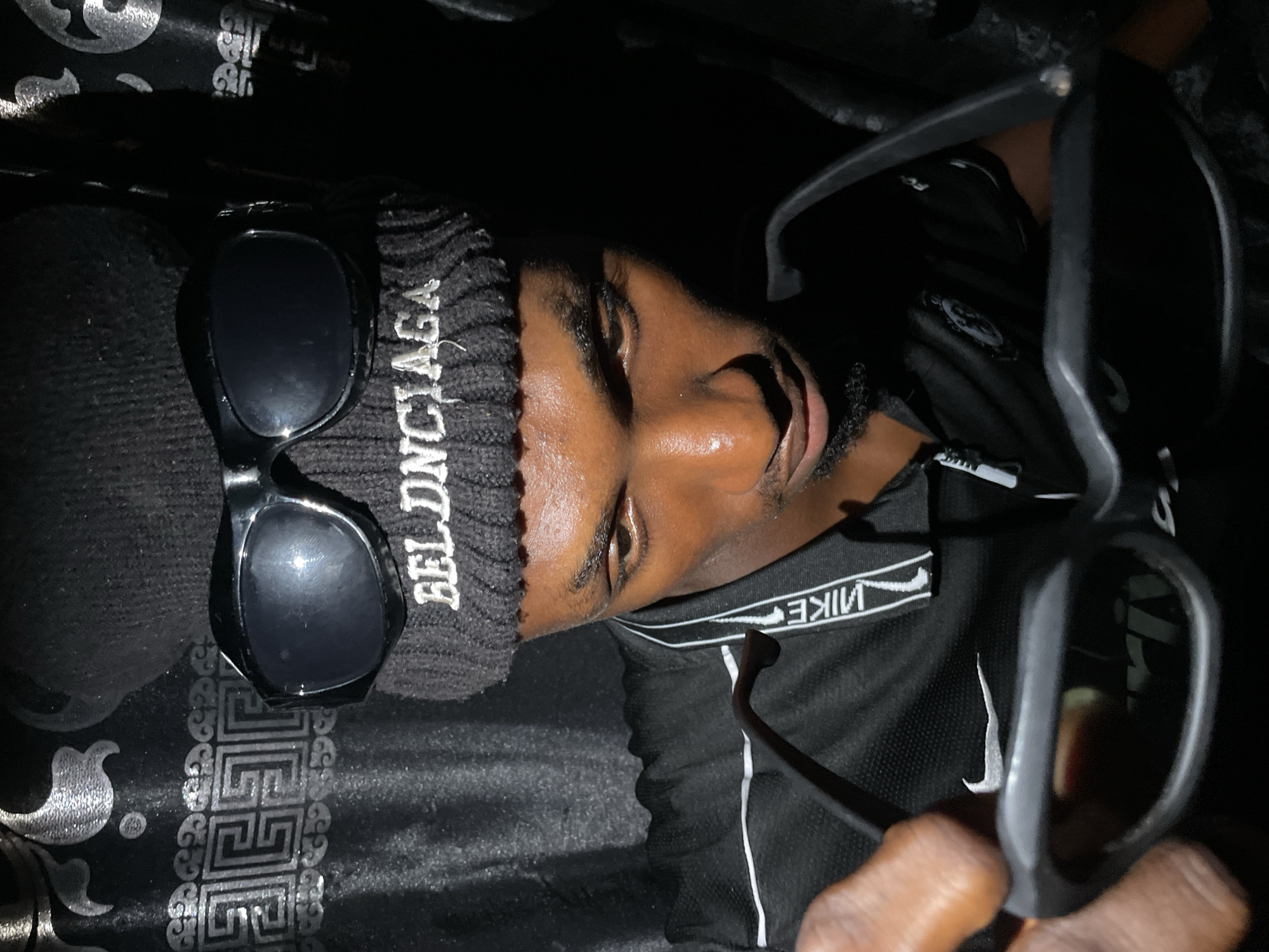 The image shows a person with dark skin wearing a black beanie with the word "BELONCIAGA" in white lettering, and a black Nike t-shirt with a white logo. The person is holding a pair of black sunglasses in their hand and another pair is lying on their head. The background is a dark, patterned fabric. The location is unknown but there is no evidence to place it in Lokoja, Nigeria.