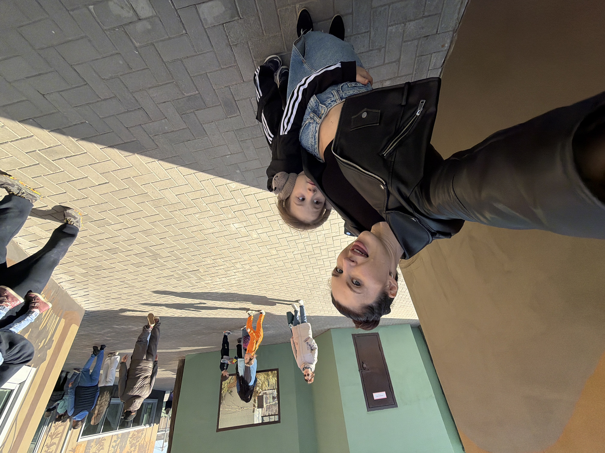 Here's a description of the image:
The image shows a tilted selfie taken by a woman in Perm, Russia. She is wearing a black leather jacket and holding a young child, who is wearing a black and white striped jacket and ear muffs. The child is looking at the camera. Behind them is a gray brick walkway. In the background, other people are walking around, and there is a building with green and brown walls. A window with a reflection of a building is visible on the green wall.