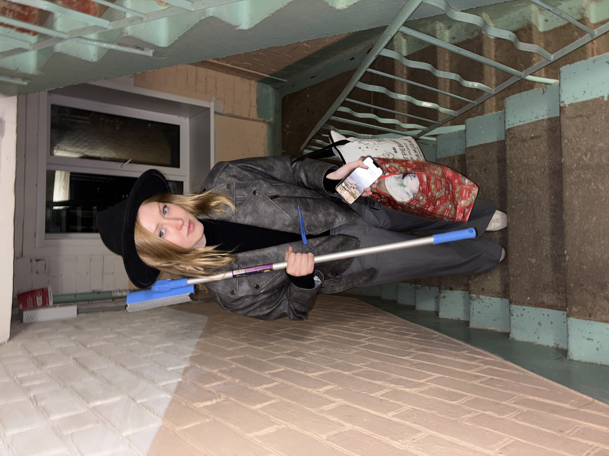 This media file, rotated 90 degrees clockwise, shows a young person standing on a utilitarian indoor staircase, likely within a building in Perm, Russia. The setting appears to be a common stairwell or a service area.
The subject, a young individual with long blonde hair, looks directly at the camera with a neutral expression. They are dressed in a black wide-brimmed hat, a black top, a grey leather-look jacket, grey trousers, and white sneakers. In their right hand, they hold an Apple iPhone displaying "2:37" and an image on its screen. Their left hand grips a blue and silver handled cleaning tool, possibly a mop or window squeegee.
Next to the person, a red patterned shopping bag, seemingly full of white contents, rests on the stairs. A white bag partially visible behind it clearly displays the Cyrillic text "ПЯТЕРОЧКА," identifying it as a bag from a Russian supermarket chain.
The environment consists of concrete stairs, with the risers and parts of the stringers painted a light blue or teal color. A light blue metal railing runs alongside the staircase. The walls exhibit a mix of painted surfaces and exposed brickwork. A window is visible in the background, and above, an industrial-style ceiling with exposed beams or joists is also painted light blue. The floor in the foreground is tiled with light brown/beige brick-patterned tiles. The artificial lighting suggests an indoor setting, and while the phone indicates "2:37," the specific time of day (AM/PM) is not explicitly clear.