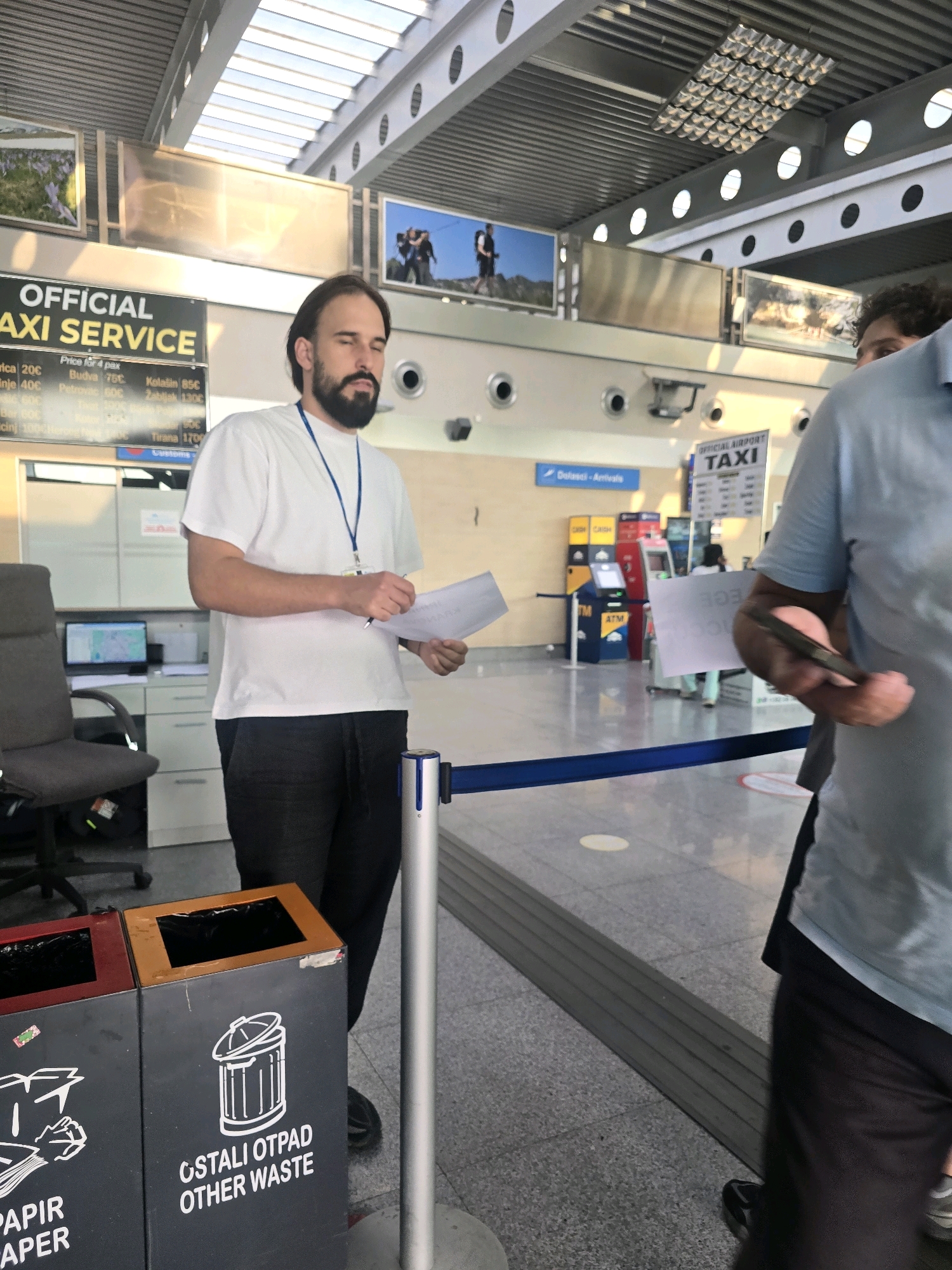 The image captures a scene likely at Podgorica Airport in Montenegro, indicated by the "OFFICIAL TAXI SERVICE" sign. In the foreground, a man with dark hair and a beard, wearing a white t-shirt and dark pants, stands with his eyes closed, holding a pen and paper. He appears to be an employee of the taxi service. To his right, another person, partially visible and wearing a light blue shirt, is holding a dark object, possibly a phone or tablet, and a piece of paper. Behind them, several ATM machines are visible, along with the "DELASCI - ARRIVALS" sign, confirming the airport setting. The environment is a well-lit indoor space with a tiled floor, a modern ceiling, and decorative pictures on the walls. Two bins for waste disposal are present in the foreground, one with a label for "PAPIR PAPER" and another for "OSTALI OTPAD OTHER WASTE." A blue rope barrier separates the areas.