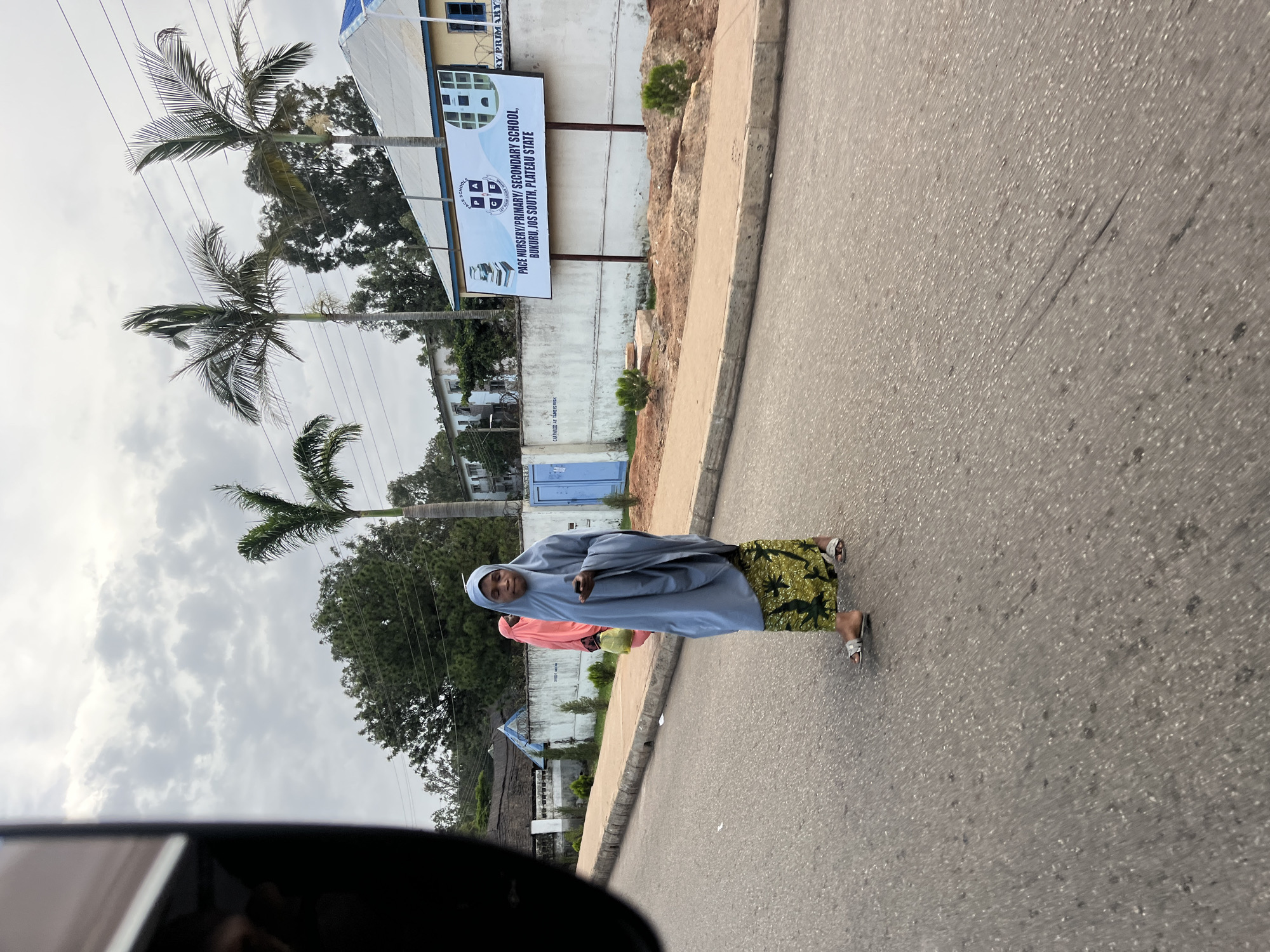 A woman in a light blue hijab and a patterned green skirt stands in the middle of a paved road, holding a smartphone in her right hand. Behind her, another person in a pink headscarf carries a yellow bag. The background shows a white wall with a sign for "PACE NURSERY/PRIMARY SECONDARY SCHOOL, BUKURU, JOS SOUTH, PLATEAU STATE," palm trees, and buildings under a cloudy sky. The road has a raised curb on the left and dirt on the right.