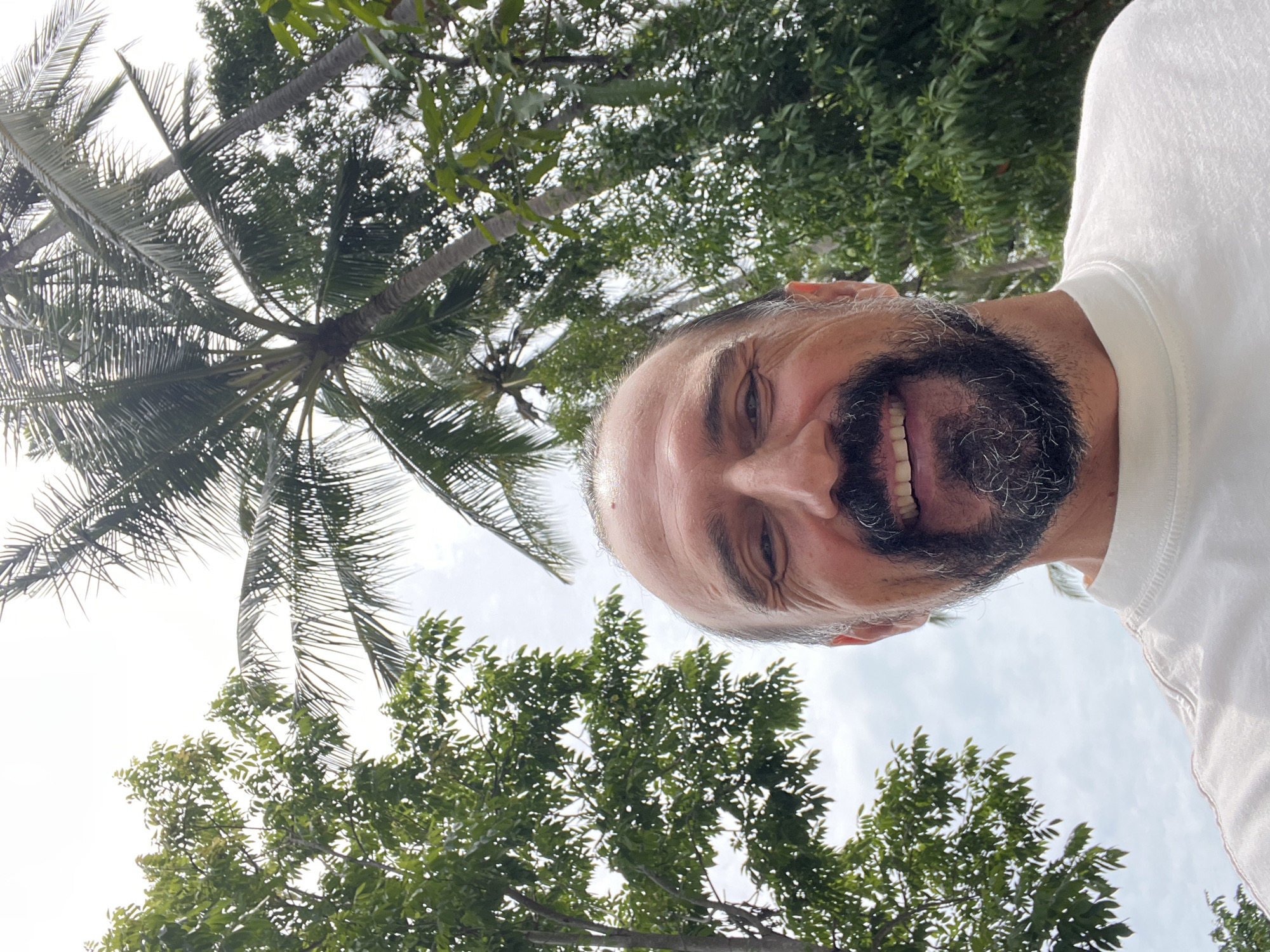 A man is looking up and smiling at the camera. He has light skin, short hair, and a gray and black beard. He's wearing a white shirt. The background features palm trees and other green trees. The scene is outdoors in a tropical environment, likely in บ้านบางรักษ์, Thailand. The sky is visible between the trees and appears slightly overcast.