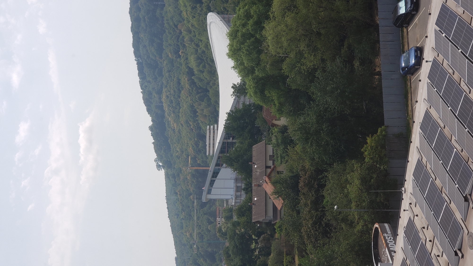 The image shows a view of the pre-construction of the Steel Arena in Prešov, Slovakia. A large, modern building with a distinctive wavy roof is under construction in the middle ground. Cranes are visible on the site, indicating ongoing construction work. In the foreground, solar panels cover the roof of a building. A blue Audi and a black car are parked on the asphalt surface behind a corrugated metal fence. Residential houses with red tile roofs are nestled among trees in front of the construction site. The background consists of a lush, tree-covered hill under a hazy sky with scattered clouds. The overall scene depicts an urban landscape with a mix of residential, commercial, and ongoing construction elements.