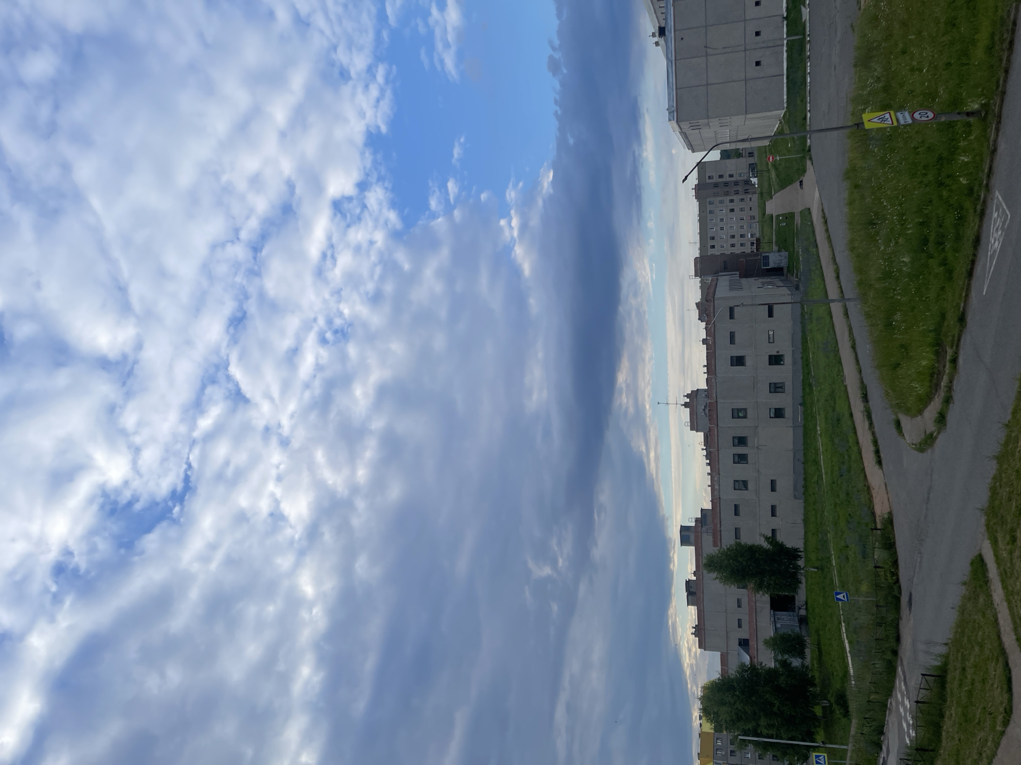 The image captures an outdoor scene, likely in the city of Markovskiy, Russia, under a partly cloudy sky. The foreground shows a gray asphalt road with a faint white marking, curving to the right. Adjacent to the road is a patch of overgrown green grass dotted with white wildflowers. Further back, another paved path, narrower than the road, winds through a grassy area.
Dominating the midground are several Soviet-era apartment buildings, characterized by their light gray facades and numerous rectangular windows. These buildings appear to be of varying heights and designs. Trees, mostly slender and dark green, are interspersed in front of the buildings, some of which have red roofs.
The background consists of more apartment buildings stretching across the horizon, suggesting a residential or industrial district. A tall light pole with a sign featuring a yellow triangle with an icon and a red circular sign with the number "20" is visible to the right. Another light pole stands near the center of the frame. The sky is a mix of bright blue patches and large, gray, and white clouds, with some darker cloud formations present. The overall impression is a somewhat overcast, possibly early evening or late afternoon view of a cityscape.