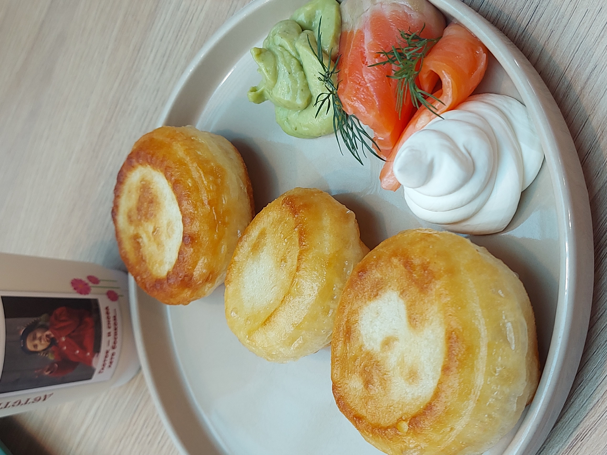 A plate is arranged on a light-colored wooden surface. The plate contains three small, golden-brown round pastries. Accompanying the pastries are dollops of green avocado spread, smoked salmon slices garnished with dill sprigs, and a swirl of white cream. A cup with a photograph on it is placed next to the plate. The cup's image includes a person wearing red clothing. The cup also has text written in a Cyrillic script. The overall image suggests a meal setting, possibly a breakfast or brunch. The Cyrillic text and cuisine may suggest the location as Saint Petersburg, Russia.