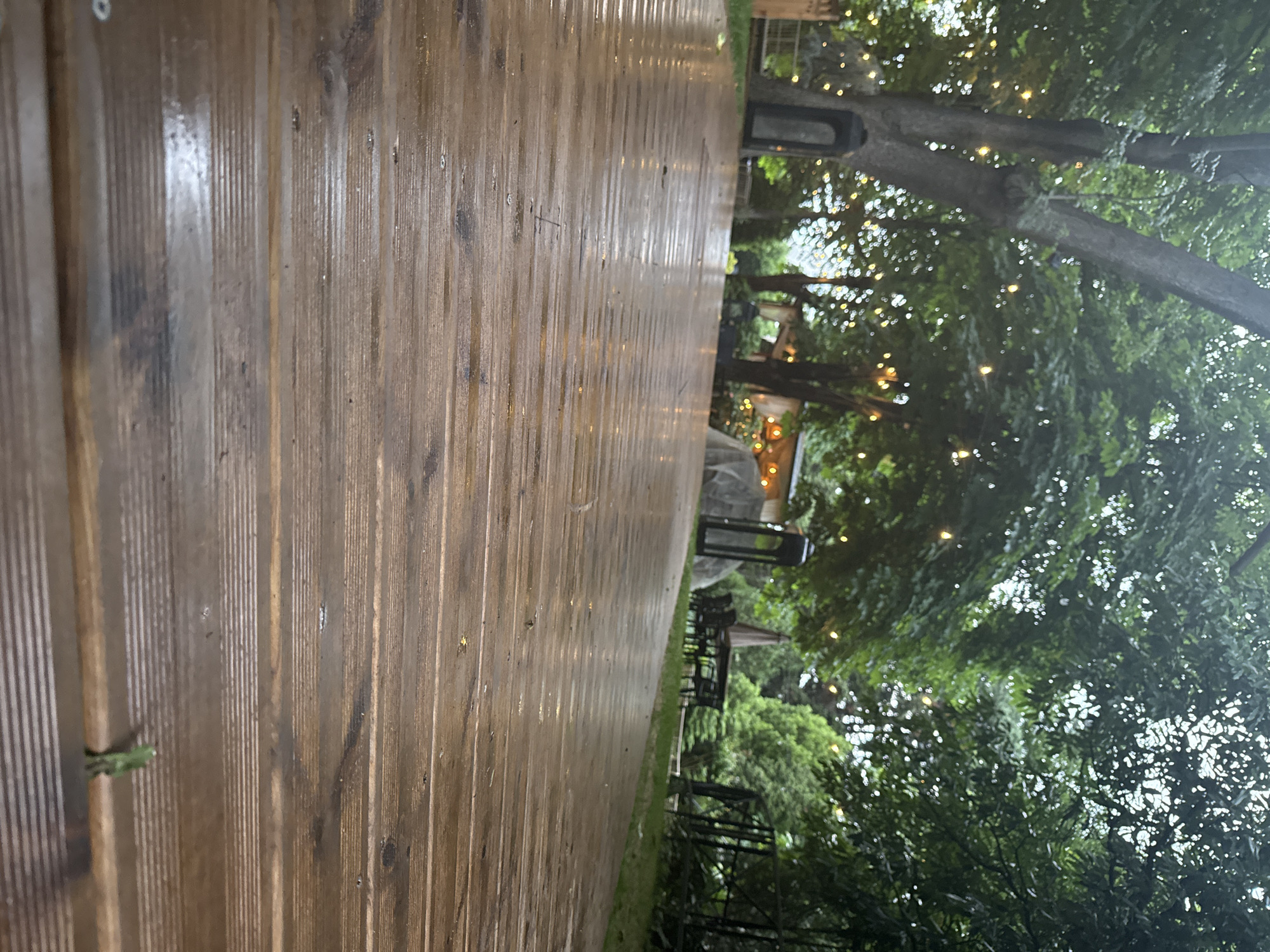The image depicts a wet wooden deck in an outdoor setting, likely a garden or patio area. The deck is made of weathered wooden planks, oriented horizontally and showing a glossy sheen due to moisture, possibly from recent rain. In the background, several large trees with lush green foliage dominate the scene. String lights are visible, hung among the trees, some illuminated, casting a warm glow.
Further in the background, various structures and objects are present. To the left, a dark, tall, lantern-like object stands near a wall. In the center, a set of outdoor dining furniture, including a table and chairs, is partially visible under a muted beige umbrella. To the right, a black metal fence or railing can be seen. The overall environment suggests a tranquil, possibly evening or dusk setting.
The image does not contain any people. The setting appears to be a private residence or a commercial establishment like a restaurant or café with an outdoor seating area. There are no discernible clues within the image to pinpoint the exact location to Varna, Bulgaria.