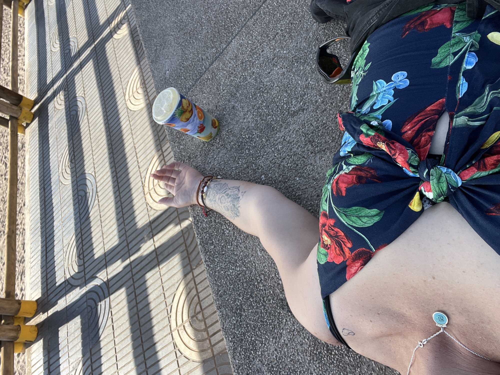 A top-down shot shows a person wearing a floral-print top knotted at the waist, lying on a patterned gray concrete surface outdoors in Arica, Chile. The person's arm, adorned with bracelets and a tattoo, extends towards a colorful disposable cup. Sunglasses and a black bag are nearby. Sunlight casts strong shadows from a railing onto the concrete surface. A silver necklace rests on the person's skin.