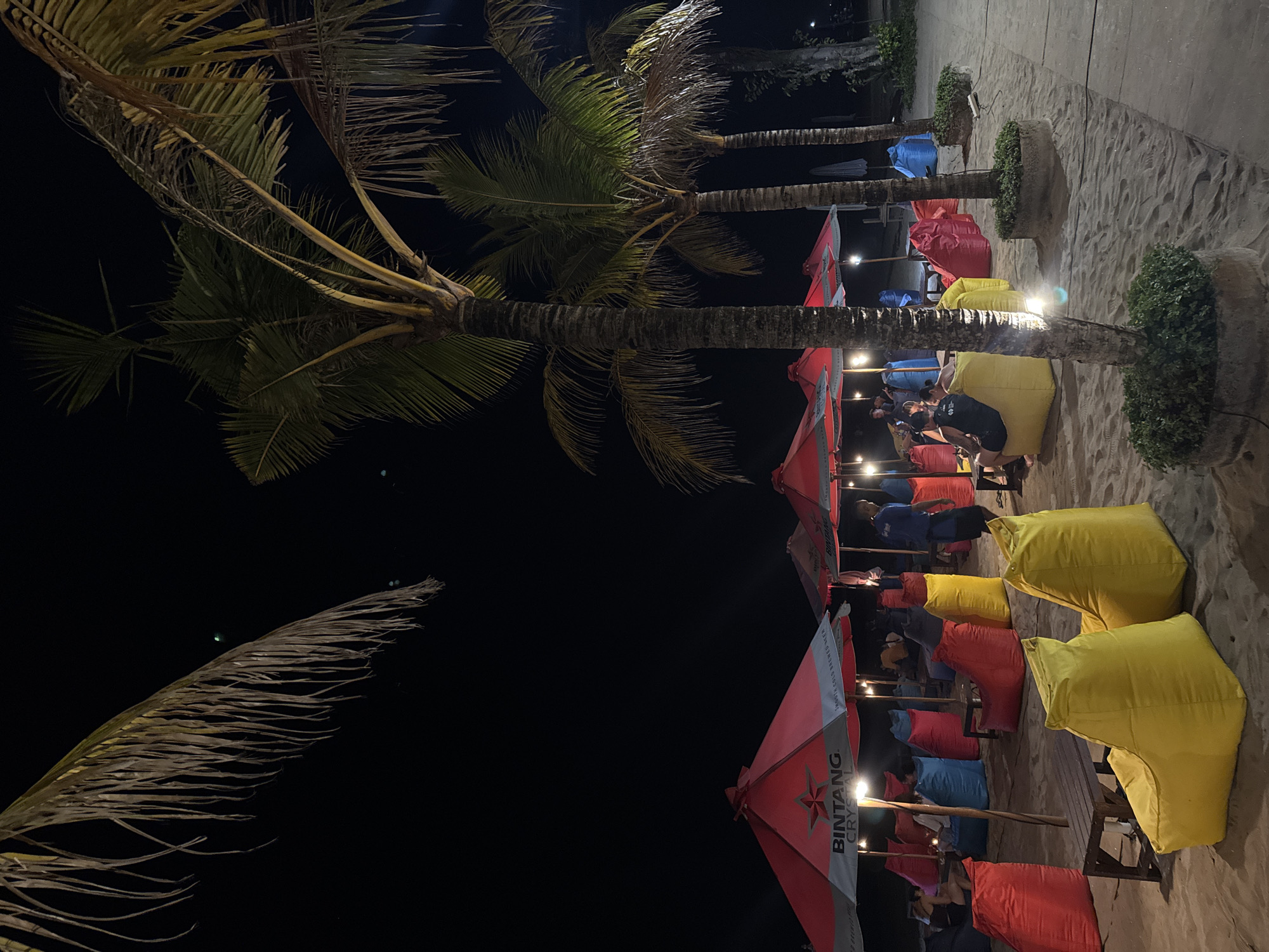 A nighttime scene on a sandy beach in Kuta, Indonesia, depicts an outdoor seating area with brightly colored beanbag chairs and red umbrellas. Several palm trees are visible, with their fronds reaching into the dark sky. People are seated in the beanbag chairs, some under the umbrellas, suggesting a relaxed atmosphere, possibly a bar or restaurant. The overall lighting is dim, with artificial lights illuminating the seating area and casting shadows from the palm trees.