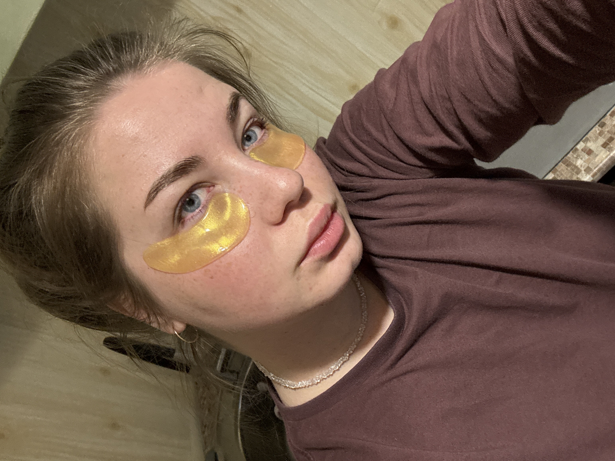A person, likely a female, is featured in a selfie. She is wearing gold-colored under-eye masks on both eyes. She has fair skin, light-colored hair, and blue eyes. She is wearing a brown long-sleeved shirt and a clear beaded necklace. A simple gold hoop earring is visible on one ear. The background is a light-colored wooden wall and a dark-colored countertop with a textured surface. There is no indication of the location being Chaikovsky, Russia.
