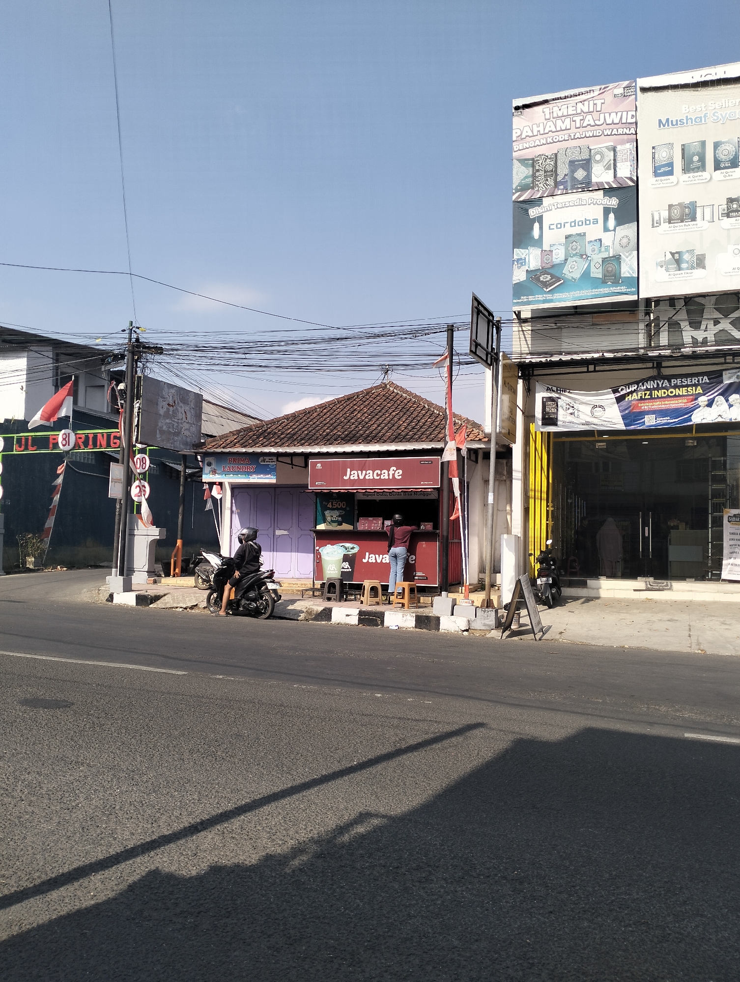 In the city of Purbalingga, Indonesia, a street scene unfolds under a clear blue sky. The image captures a row of buildings along a road. On the left, a building with a dark facade features a green sign with the text "JL. F. RING 81". Next to it, a smaller structure with a reddish-brown roof is labeled "Javacafé". A person on a black motorcycle is parked in front of the Javacafé. A second person is standing on a stool behind the counter of the Javacafé, possibly serving a customer. Further to the right, a larger building with a glass facade displays advertisements, including one that reads "GURANNYA PESERTA HAFIZ INDONESIA". The street in the foreground is asphalt, and shadows from overhead wires and a signpost are cast across it. The overall impression is a typical commercial street in a town.