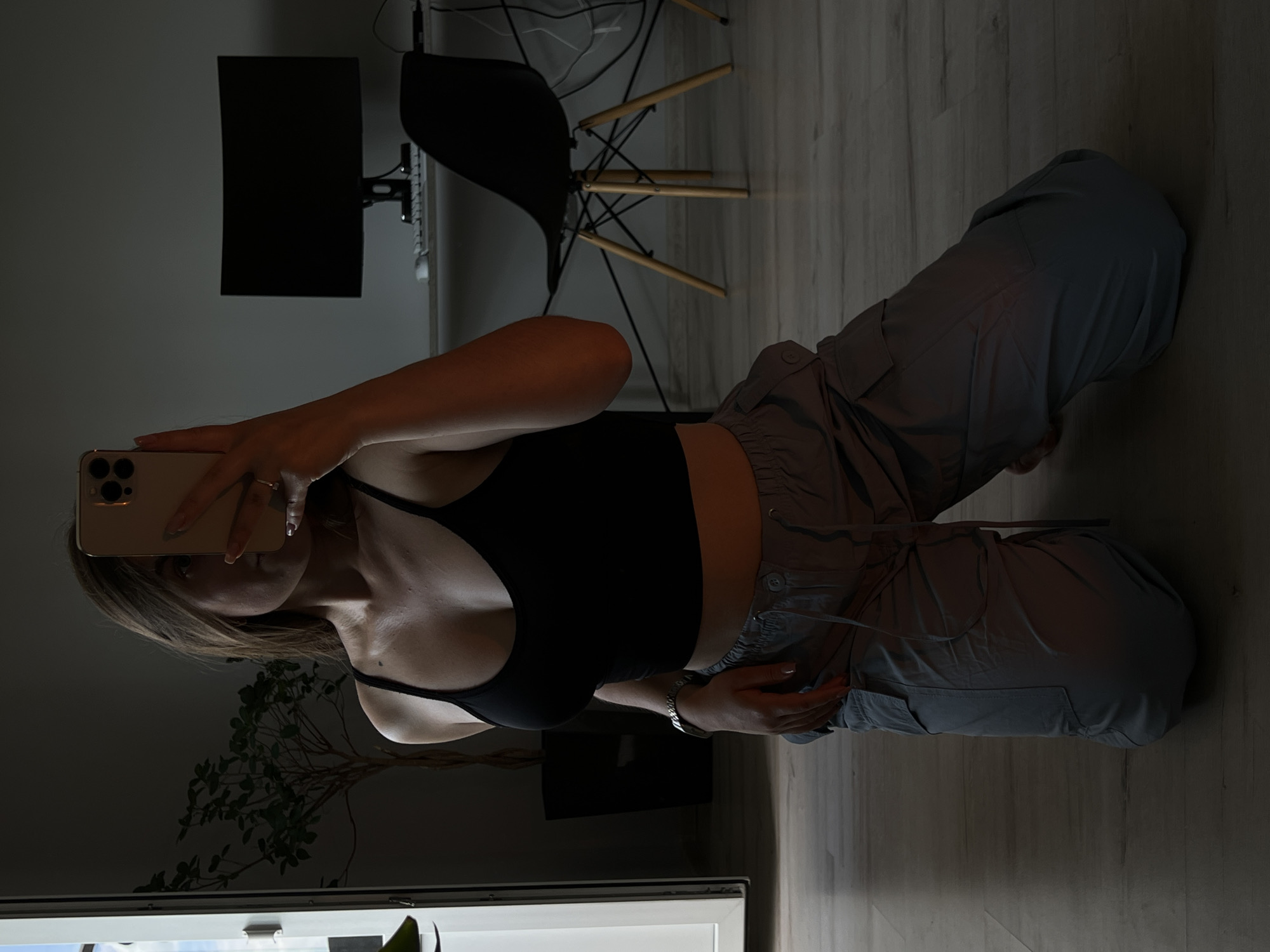The image shows a young woman kneeling on a wooden floor, holding a smartphone to take a selfie. She is wearing a black crop top and grey cargo pants. She has long brown hair and is looking at her phone. The background shows a room with a desk, a computer monitor, a chair, and a plant. The room is dimly lit, with light coming from a window on the left side of the image. This scene appears to be an indoor residential setting, likely a home office or bedroom, and there is no indication of it being in Ternopil, Ukraine.