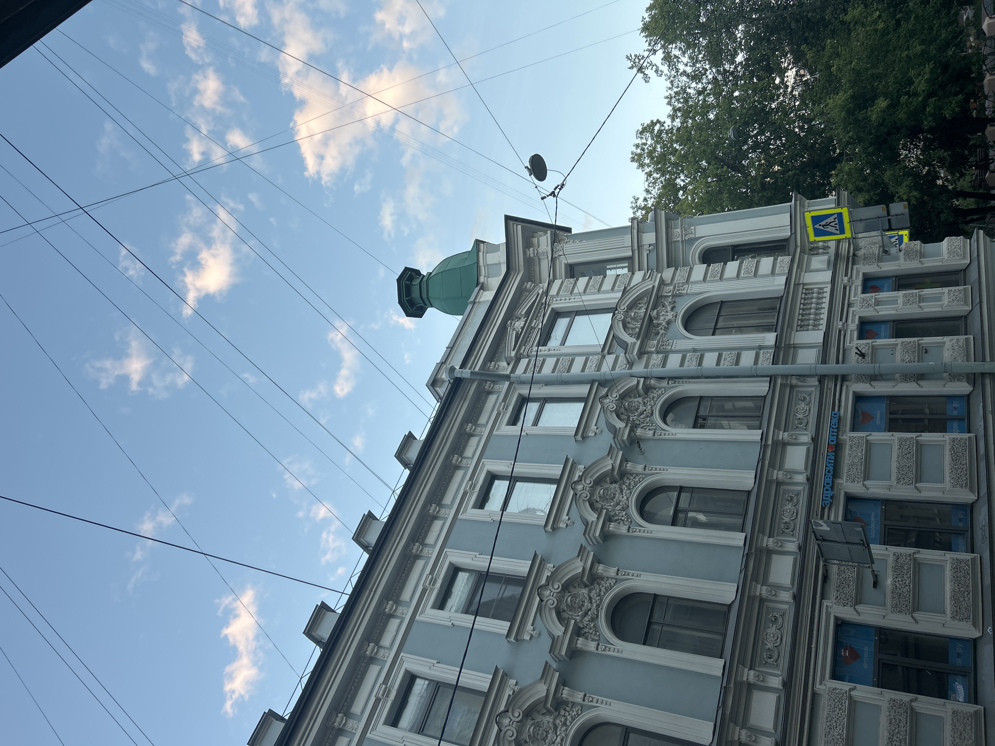 The image shows the facade of a light blue, ornate building with white trim, viewed from a low angle looking upwards. The building has multiple floors with numerous windows, some arched and some rectangular, adorned with decorative moldings. A prominent green dome tops a section of the building. Overhead, a network of power lines crisscrosses the bright blue sky, which is dotted with fluffy white clouds. To the right, a large green tree with dense foliage overhangs the building, and a yellow and blue pedestrian crossing sign is visible. The lighting suggests it is daytime, likely late morning or afternoon, with the sun casting gentle shadows. The scene appears to be an urban setting, possibly a street in a historic district, given the architecture. There is no visible activity or people in the frame. On the building, text is visible on blue signs in some of the lower windows, appearing to be Cyrillic script that reads "ЗдравСити Оптика" and "ЗдравСити Оптика", suggesting a pharmacy or optical store.