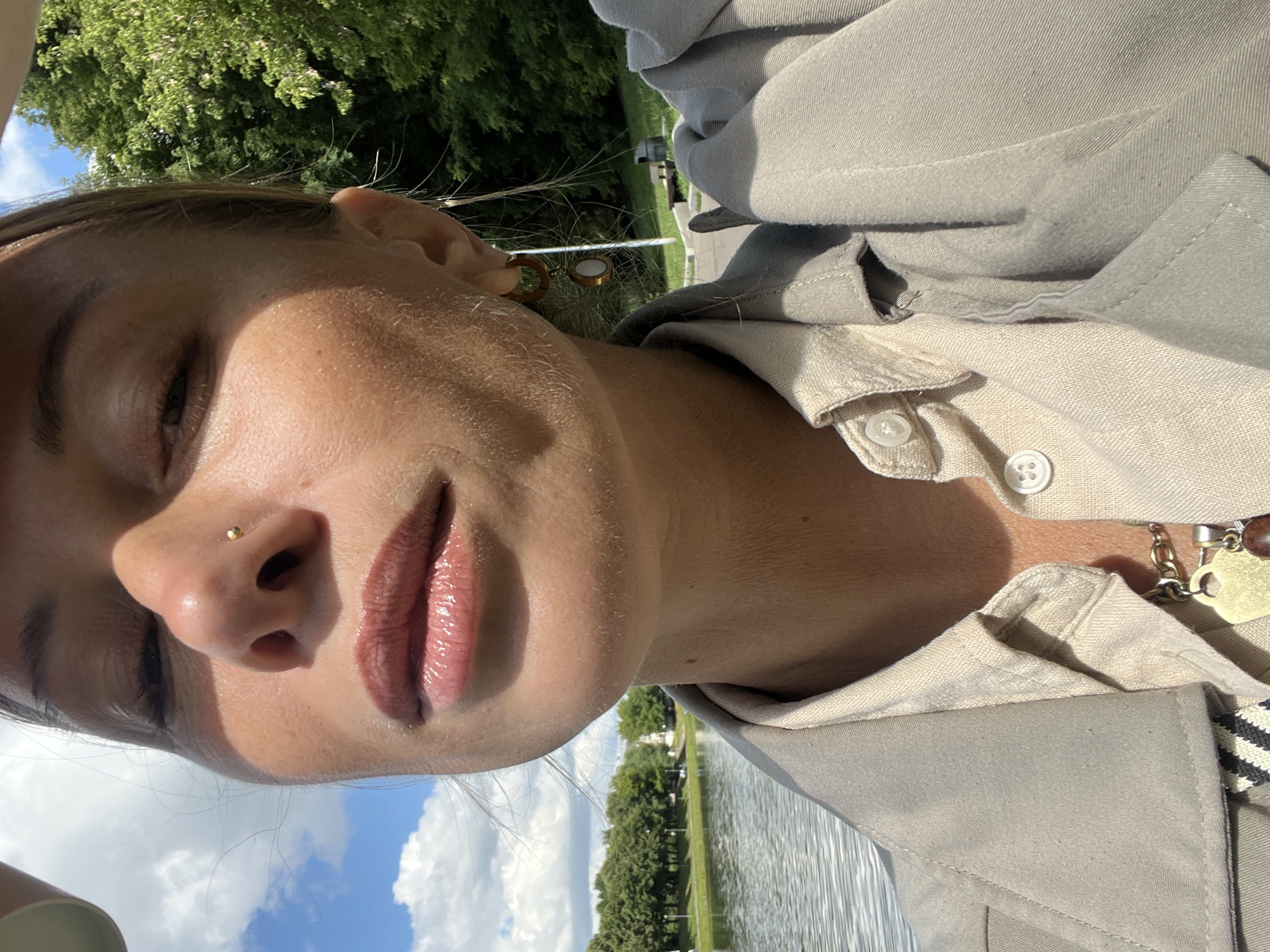 This is a close-up, vertically oriented photograph of a person's face and upper body, taken outdoors on a sunny day. The person's face is turned towards the camera, with their eyes closed and their lips slightly parted, suggesting a relaxed or contemplative mood. A subtle nose stud is visible. The person is wearing a light-colored collared shirt or jacket, with visible buttons. A necklace with a pendant is also visible around their neck.
The background features a body of water, likely a river or lake, with trees and greenery along the bank. The sky is visible with some white clouds. The lighting suggests it is daytime, likely late morning or afternoon, with the sun casting shadows and highlighting the person's skin. The overall setting appears to be a park or waterfront area. There is no visible text in the image. The context provided indicates the location as Moscow, Russia.