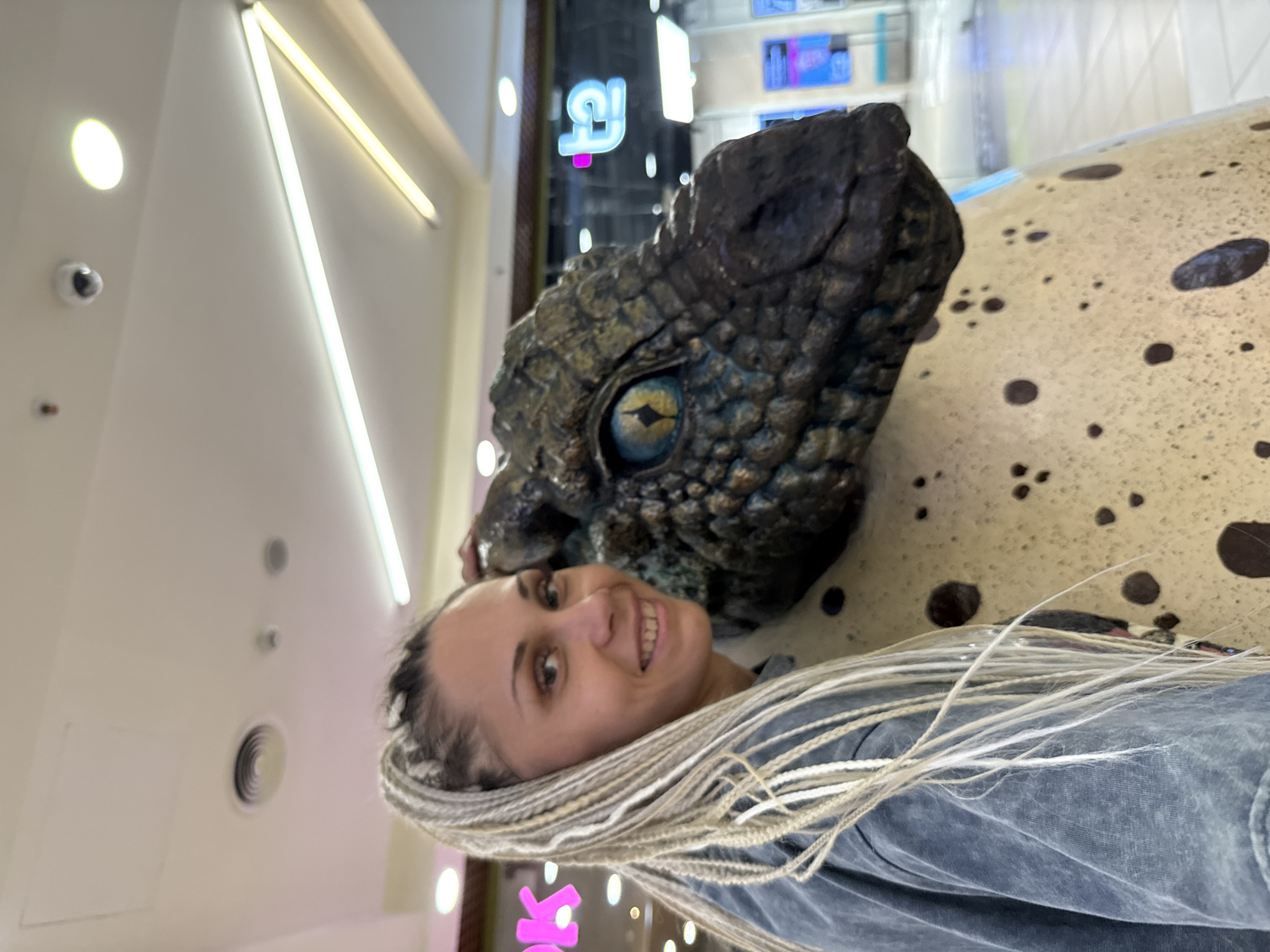 A woman with blonde braids is posing with a dinosaur statue in what appears to be an indoor location, possibly a museum or shopping mall, given the lighting and the backdrop. The dinosaur statue is a close-up, with its head and eye visible, and it sits on a spotted, egg-like surface. The woman is smiling and looking at the camera, with her braids cascading down. The lighting is bright, suggesting it's daytime, and the setting is indoors. There are no obvious weather cues. A neon sign with Cyrillic letters, "Арк" (Ark), is visible in the background, which might indicate the location is in Arkhangelsk, Russia.