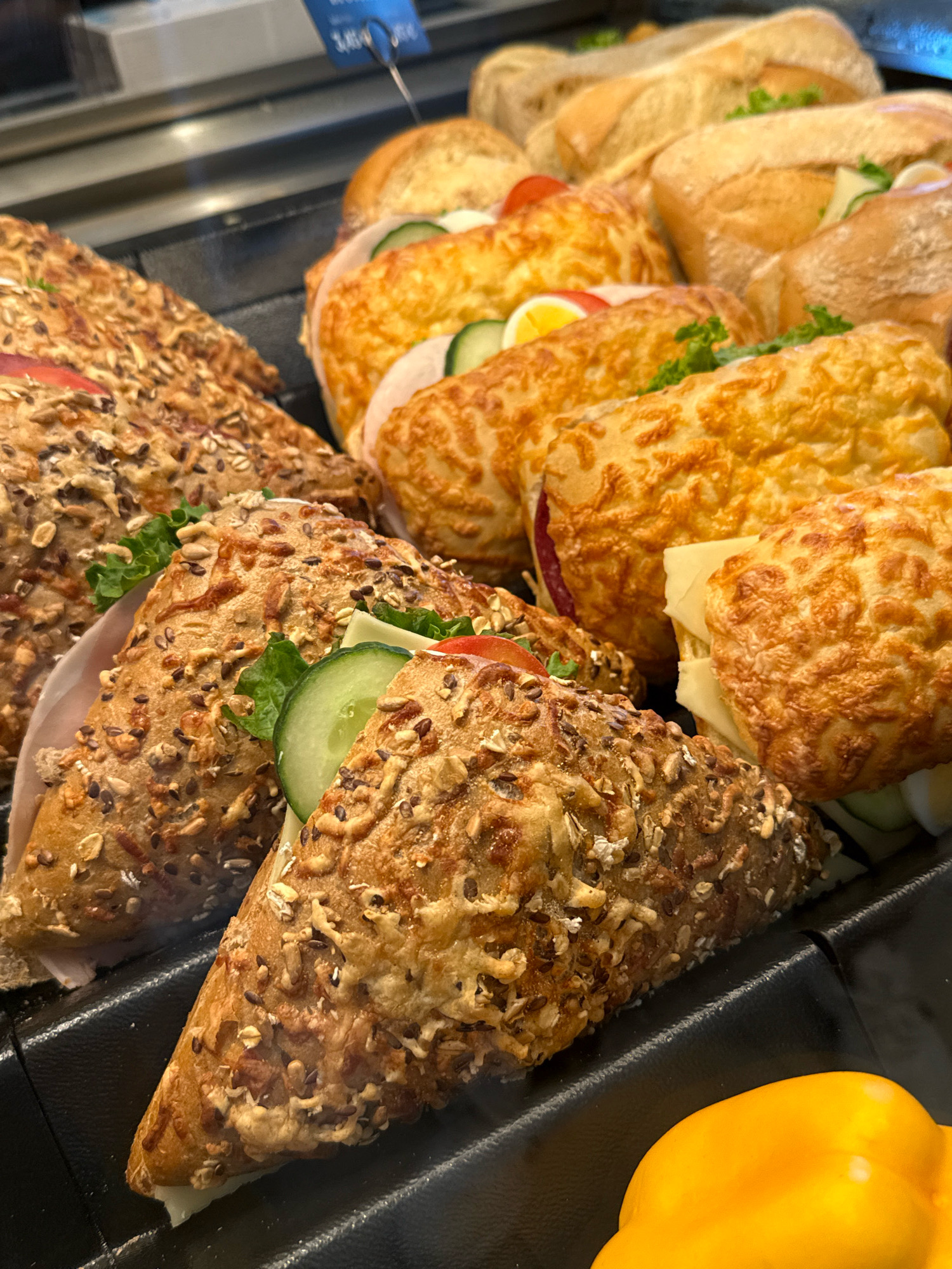 The image displays a close-up view of a display case filled with a variety of sandwiches. In the foreground, two triangular sandwiches with whole grain bread and visible seeds and grains are prominent. These sandwiches appear to be filled with lettuce, cucumber slices, and possibly ham or salami. Behind them, several other sandwiches are visible, some with golden-brown crusts that suggest they may be toasted or baked with cheese. These include round rolls and larger rectangular sandwiches. The overall setting suggests a bakery or a food service establishment. A yellow bell pepper is partially visible in the lower right corner. The lighting is bright, illuminating the food items and creating shadows. The background is blurred, but appears to be the interior of a commercial space. There are no people visible in the image. The image was taken at a bakery in Herzberg, Germany.