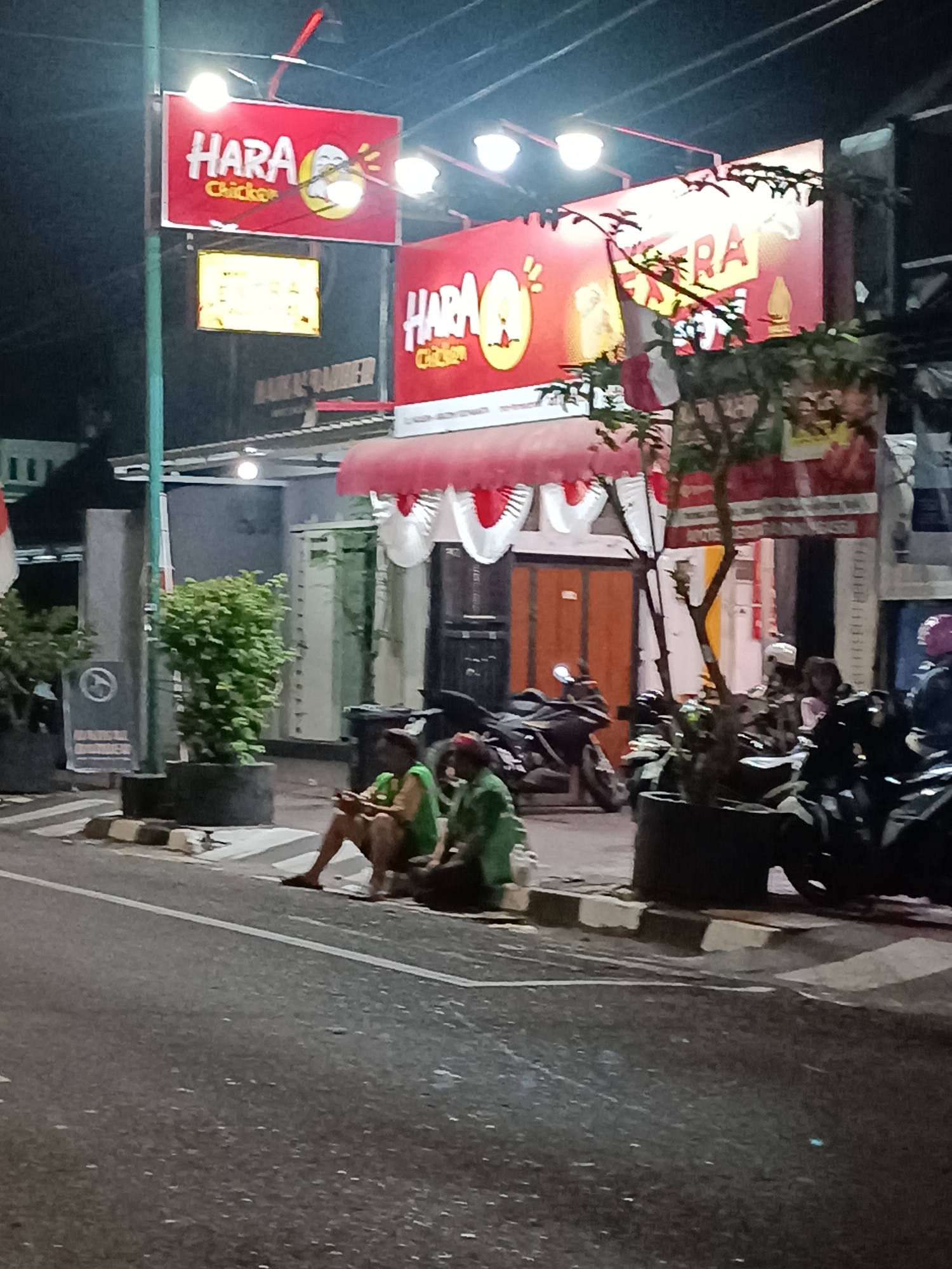 The image shows a street scene at night in the city of Yogyakarta, Indonesia, likely in an area with food establishments. The primary subject is a brightly lit red sign that reads "HARA Chicken" in white lettering, with a cartoon chicken graphic. To the right, another similar sign is partially visible. Below these signs, a covered outdoor seating area is visible with red and white decorations. A group of people are sitting on the curb in front of the establishment, some appearing to be using their phones. Several motorcycles are parked on the street. The overall atmosphere suggests a casual, late-night dining or gathering spot.
