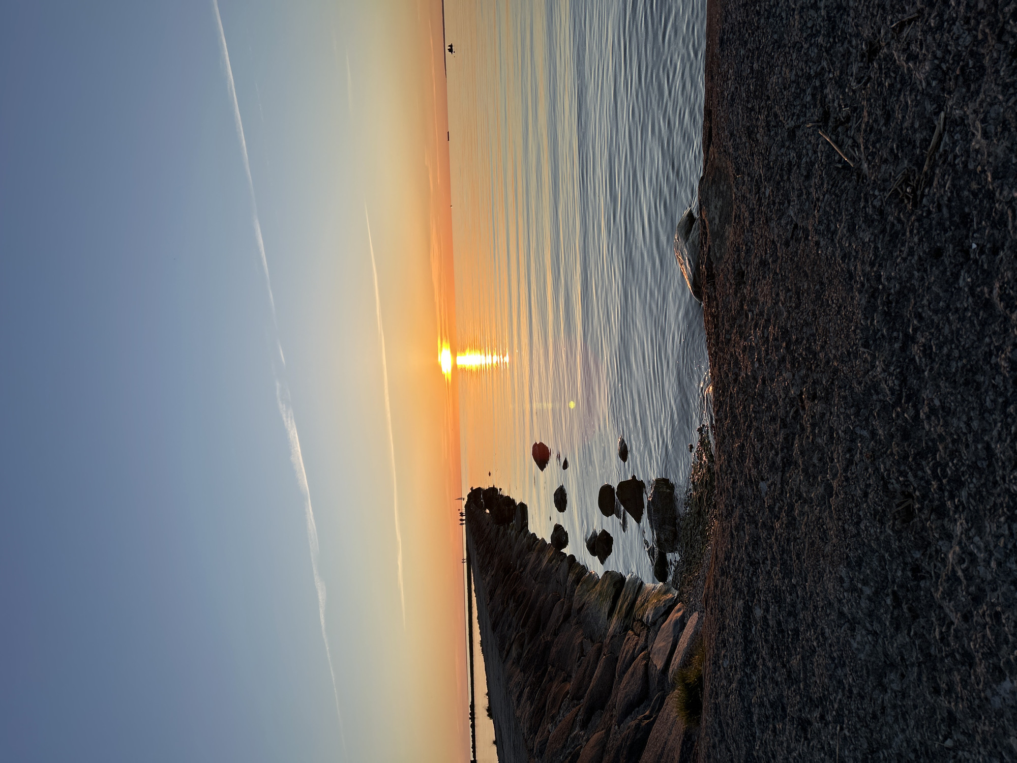 The image depicts a sunset over a calm body of water, likely the Baltic Sea, viewed from a low angle on a pebbly shoreline. In the foreground, a dark, granular, pebbly or gravelly beach occupies the bottom third, exhibiting subtle undulations and scattered small pieces of wood or debris.
Dominating the mid-ground is a robust, angular stone breakwater or pier, constructed from large, dark, irregular rocks. It extends from the lower-left corner, curves gently rightward, and then proceeds diagonally towards the sun, gradually receding into the distance. A flat path is visible along the top of the breakwater. Approximately four to five small, silhouetted figures are discernible walking or standing on this path in the mid-distance. Further along the breakwater, towards the sun, a distinct white triangular object, possibly a small sailboat or a marker, stands near one of the figures.
The body of water, smooth with minimal ripples, occupies the right and central portions of the image. It displays a reflective surface, mirroring the vibrant orange and yellow hues of the setting sun, which casts a bright, elongated pillar of light directly towards the viewer. The sun is a distinct, intense orange orb positioned just above the horizon, slightly to the right of the image's vertical center.
On the far-right horizon, two small, dark silhouettes, possibly individuals in a small boat or on a paddleboard, are visible. Further out, in the main expanse of the water, several larger, dark, rectangular forms, indicative of distant cargo ships or vessels, are present.
The sky, comprising the upper half of the image, transitions from a clear, light blue at the zenith to a warm, soft orange-yellow near the horizon, blending with the sunset's glow. Two distinct, thin white contrails are visible: one diagonally across the upper-left quadrant and another, less pronounced, in the upper-right.
The scene's features, particularly the stone breakwater and the general coastal landscape, are consistent with several coastal areas in Tallinn, Estonia. Based on the prominent stone breakwater and calm sea, this location strongly resembles the waterfront along the Rocca al Mare promenade or a similar coastal stretch within Tallinn, Estonia, which features extensive stone breakwaters extending into the sea.