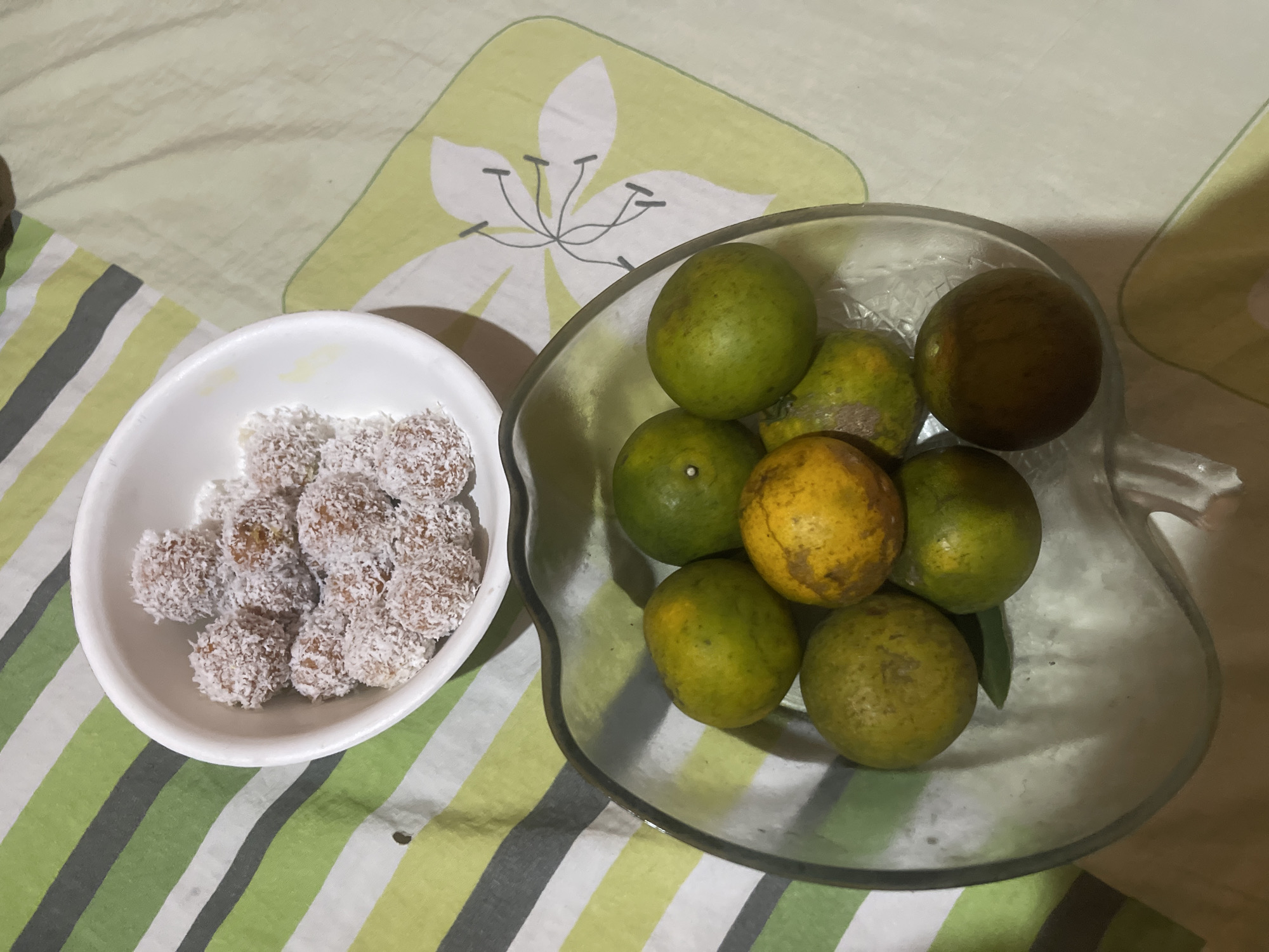 A close-up, top-down shot shows a bowl of small, round candies coated in shredded coconut next to a clear glass bowl filled with tangerines on a striped fabric surface. The candies, appearing to be about an inch in diameter, are clustered in a white bowl. The tangerines, varying in color from green to yellow and some with browned spots, are arranged in the clear bowl, which has a decorative, wavy rim. The fabric beneath has a pattern of green, white, and gray stripes on the left, transitioning to a lighter green fabric with a stylized white flower design on the right. The image is taken from an overhead perspective. This scene is located in Anggaswangi, Indonesia.