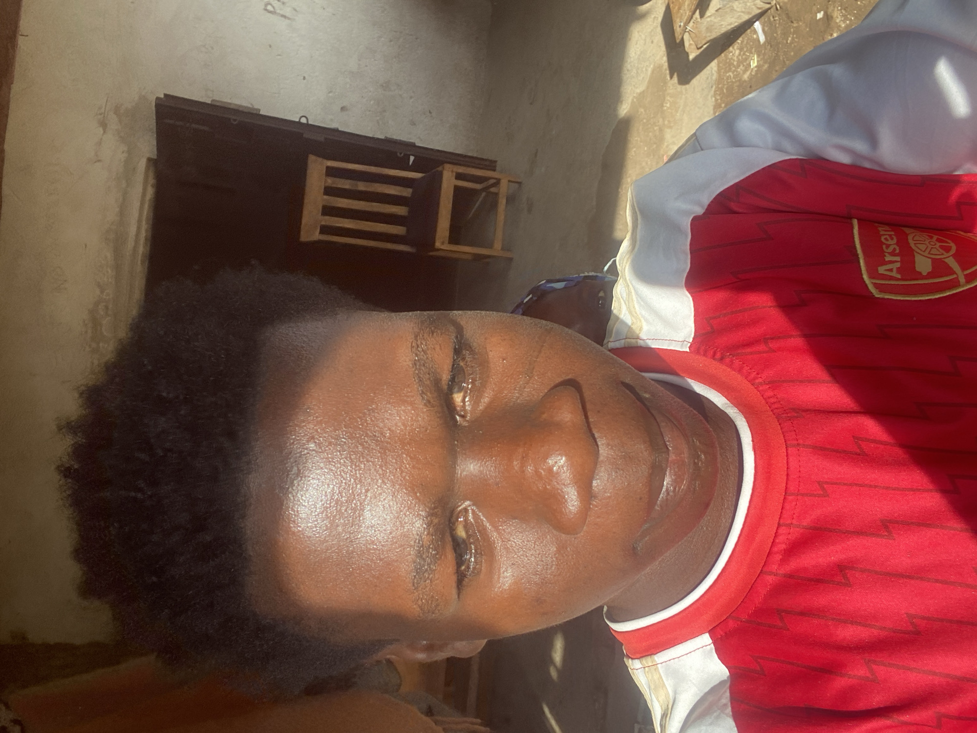 A close-up selfie of a young Black male is shown, facing forward. The individual has short, dark, curly hair and a dark complexion with visible shine on the skin due to sweat or oil. Their eyes are open and looking slightly downwards, with visible reflections. The person is wearing a red and white t-shirt with a crest on the left side of the chest. The background appears to be an indoor or semi-outdoor setting with a rough wall and a wooden chair visible. Sunlight casts a strong shadow across the subject's face and t-shirt. The overall impression is that the photo was taken during the daytime, possibly in a residential area of Mararaba, Nigeria.