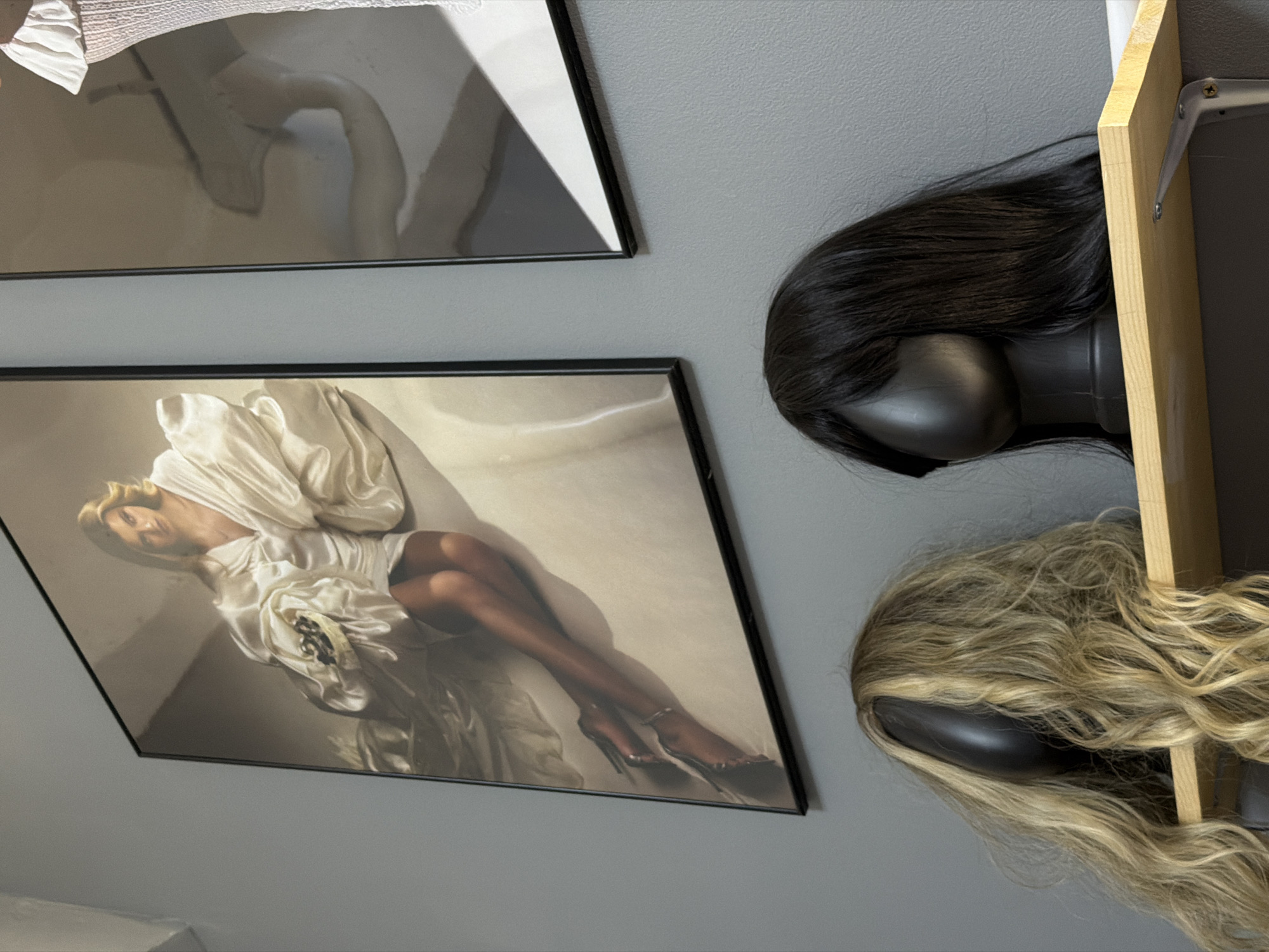 The image shows a section of a gray wall adorned with two framed pictures. The upper picture features a monochromatic image, while the lower one displays a woman in a white dress, lying in a light-toned setting. Next to the frames, a wooden shelf is mounted, holding two mannequin heads with wigs; one black and one blonde. The wigs are cascading off the shelf. The scene appears to be indoors. There is no evidence to support that this image was taken in Kazan, Russia.