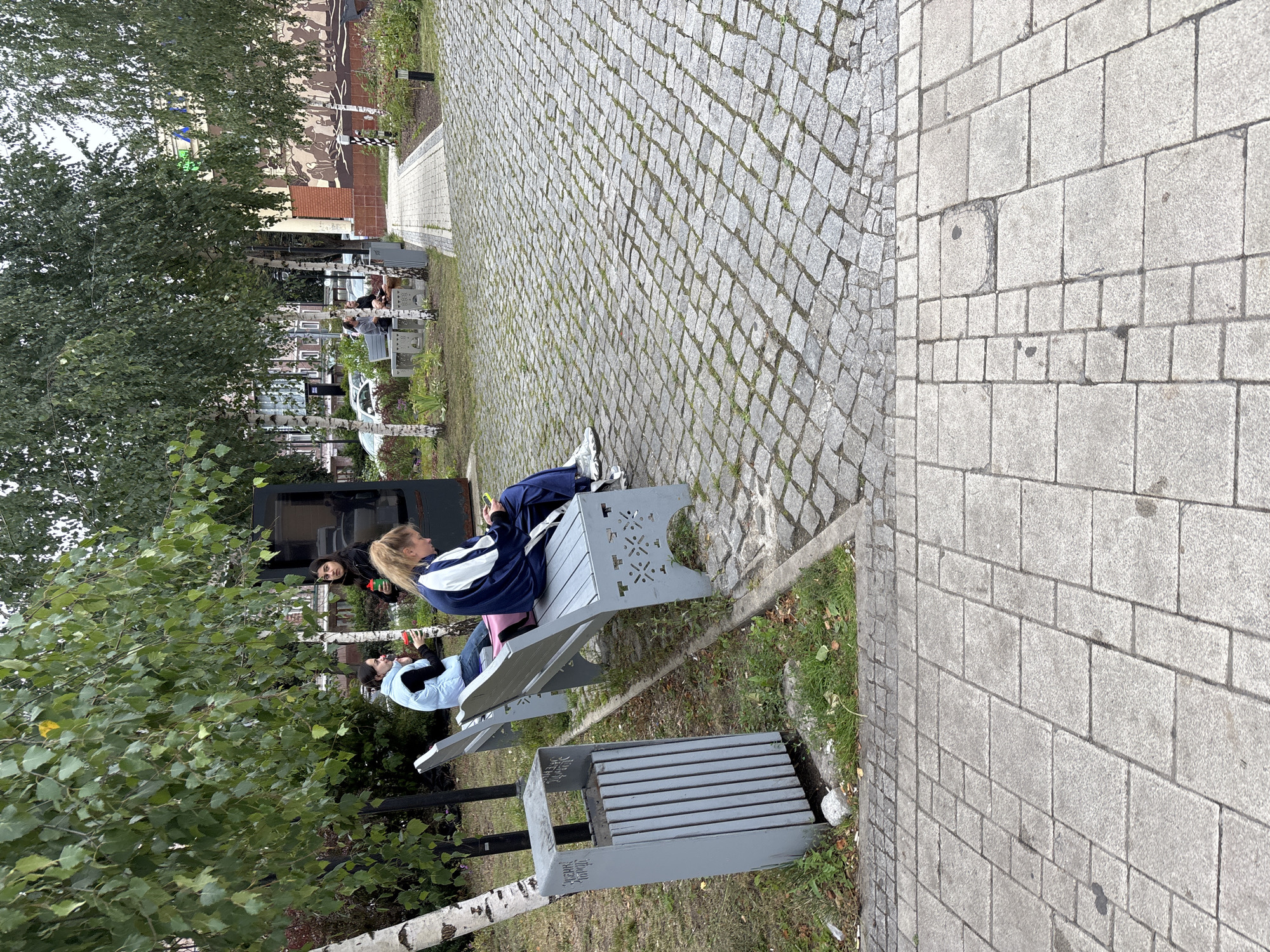 The image shows an outdoor scene in a park-like setting with paved walkways and benches. Three young women are visible. One woman, wearing a light blue jacket and jeans, sits on a grey bench, holding a red cup. Another woman, in a dark jacket, stands behind her, also holding a red cup. A third woman, wearing a blue and white tracksuit, sits on another bench, looking at her phone. A grey trash bin is located near the benches. In the background, there are trees, buildings, and cars. The pavement is made of cobblestones and large rectangular stone slabs. The overall impression is of a public space, possibly a park or a square in Zvenigorod, Russia, where people are relaxing outdoors.