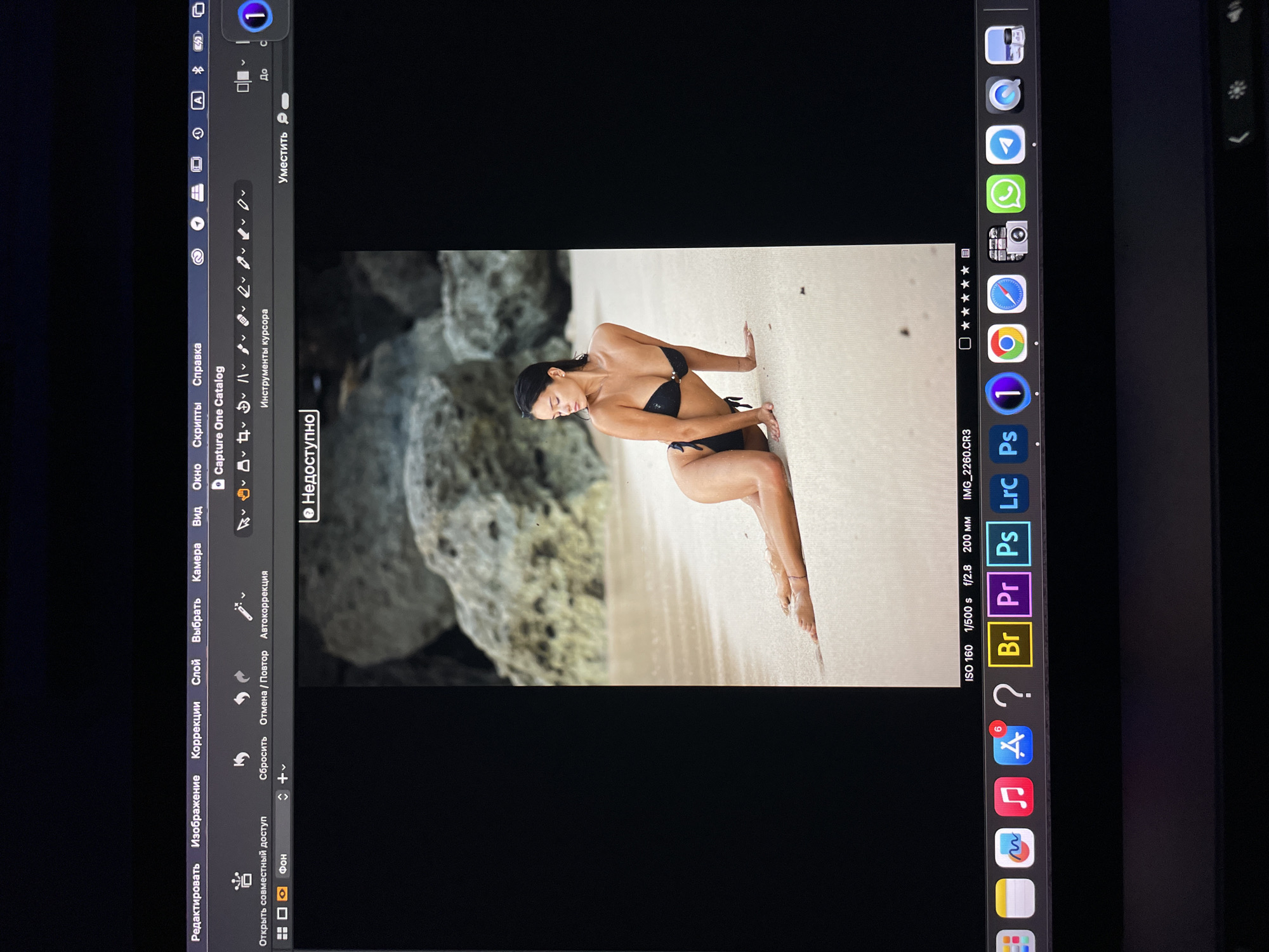 A screen capture shows the Capture One editing software open on a Mac device. The software is displaying a photo of a woman in a black bikini posing on a sandy beach near a rock formation. The Macbook screen also displays the MacOS dock with icons for Safari, Chrome, Telegram, Whatsapp, a camera application, and other Adobe Creative Suite applications like Photoshop, Lightroom, Premiere Pro, and Bridge. The top menu bar displays options like File, Edit, Image, Adjustments, Layer, Select, Camera, View, Window, Script, and Help. The photo settings are visible, including the ISO, shutter speed, aperture, and focal length. The location is not evident in the image.