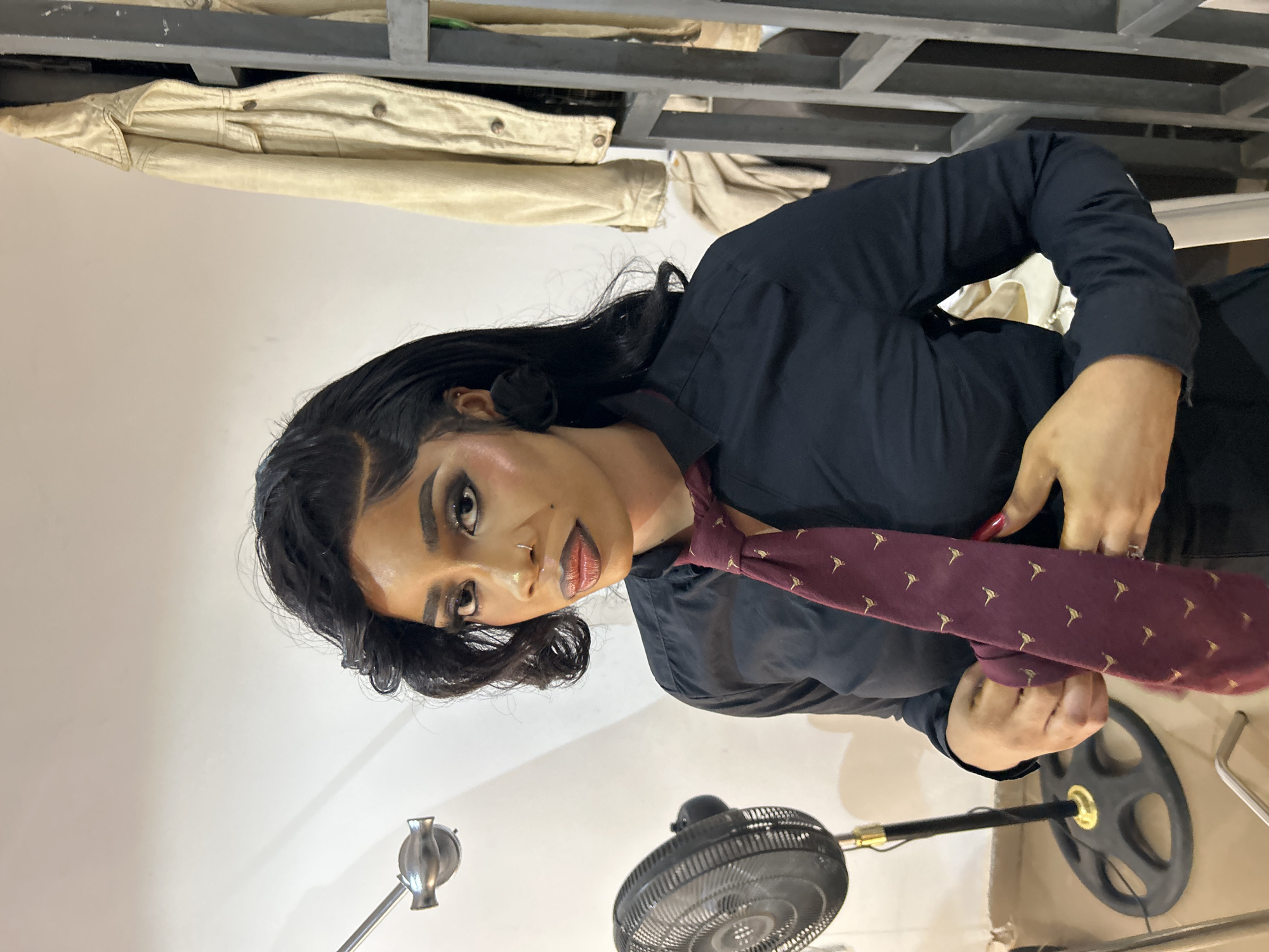The image depicts a woman wearing a dark-colored shirt and a patterned maroon tie. She is holding the tie with her hands. A fan and a floor lamp are visible in the foreground. Behind her are clothing racks with garments hanging. The setting appears to be indoors. There is no specific information provided in the image to indicate that the location is in Jos, Nigeria.
