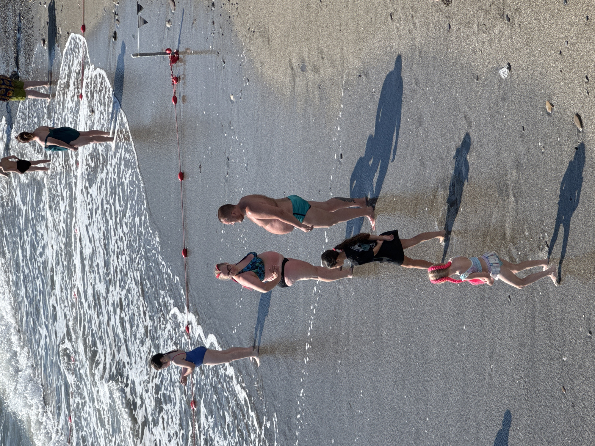 This image captures a group of people on a sandy beach in Sochi, Russia. The scene appears to be during the daytime, under sunny conditions, with gentle waves lapping at the shore. The people are dressed in swimwear and are positioned in a line along the edge of the water. Some are standing, while others appear to be in a plank or push-up position. The overall mood seems relaxed and active, suggesting a leisure activity or exercise routine.
Among the subjects, there are adults and children. One woman is wearing a green swimsuit, and another a black bikini with a patterned top. A man in blue swim trunks is visible, and a child with bright pink braids is in the foreground. There's also a woman in a blue swimsuit and a girl in a black dress.
Notable details include the shadows cast on the sand, indicating the sun's position. Red buoys on a rope are strung out along the shoreline, possibly marking a boundary or an activity area. The sand is a mix of light and dark grains, with some scattered pebbles. The water is clear with foamy whitecaps.
There is no visible text in the image. The activity appears to be a group exercise or a playful interaction with the water. The combination of sun, sand, and sea points to a typical beach day in a resort location like Sochi.