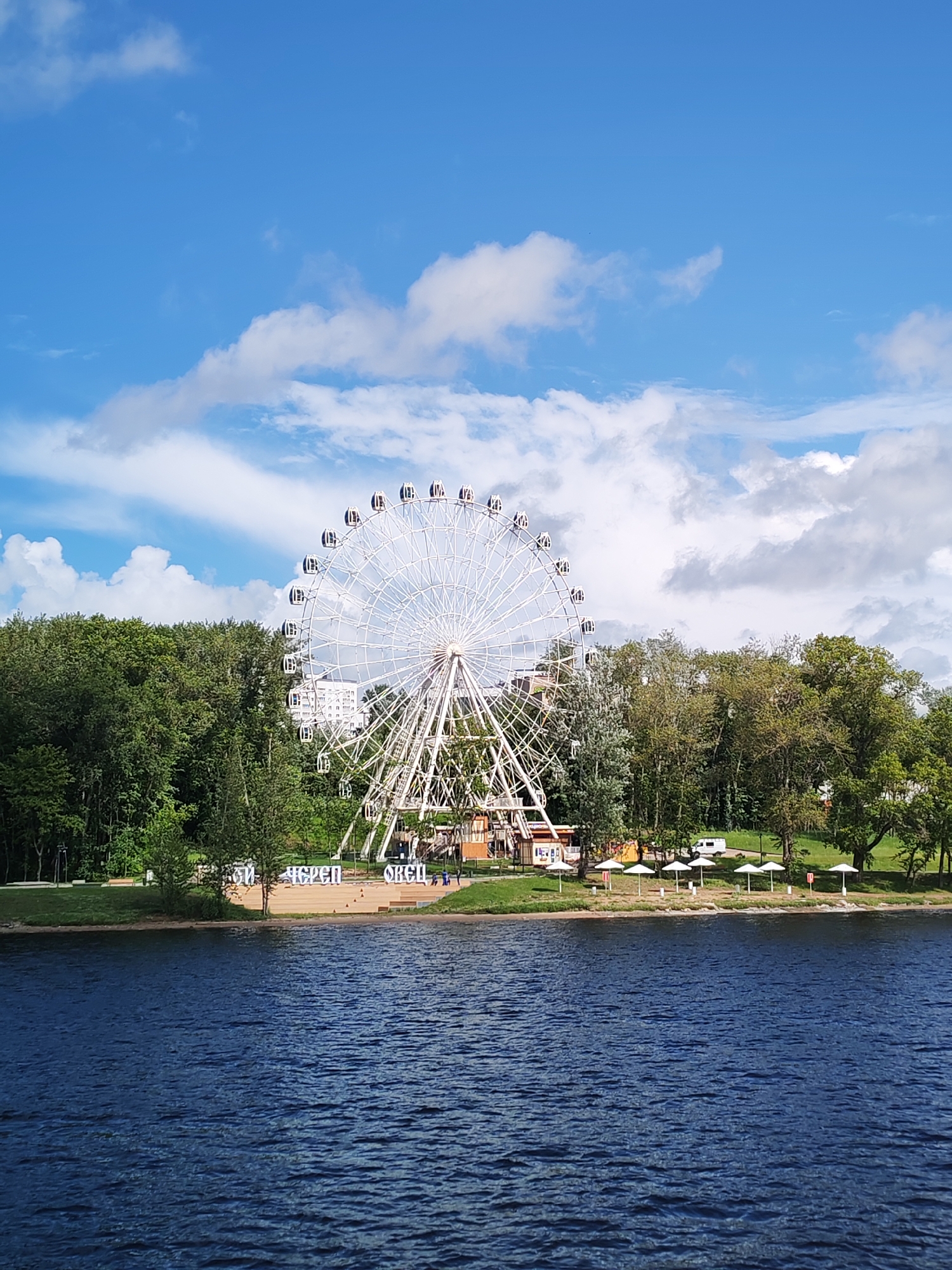 The image displays a large white Ferris wheel situated behind a line of trees and a body of water. The Ferris wheel is the dominant structure, featuring numerous cabins suspended from its spokes. In the background, several buildings are visible, including what appears to be a multi-story apartment complex. The foreground is occupied by a wide, dark blue body of water with visible ripples. Along the shoreline, a sandy area transitions into a grassy park with trees. Several white umbrellas are set up on the grassy area, and some white lettering is visible on a raised structure behind the umbrellas, which translates to "CHEREPOVETS CITY". There are also a few small, dark figures of people visible near the Ferris wheel. The sky is blue with scattered white clouds. The scene is set in the city of Cherepovets, Russia, likely in a park or recreational area adjacent to a river or lake.
