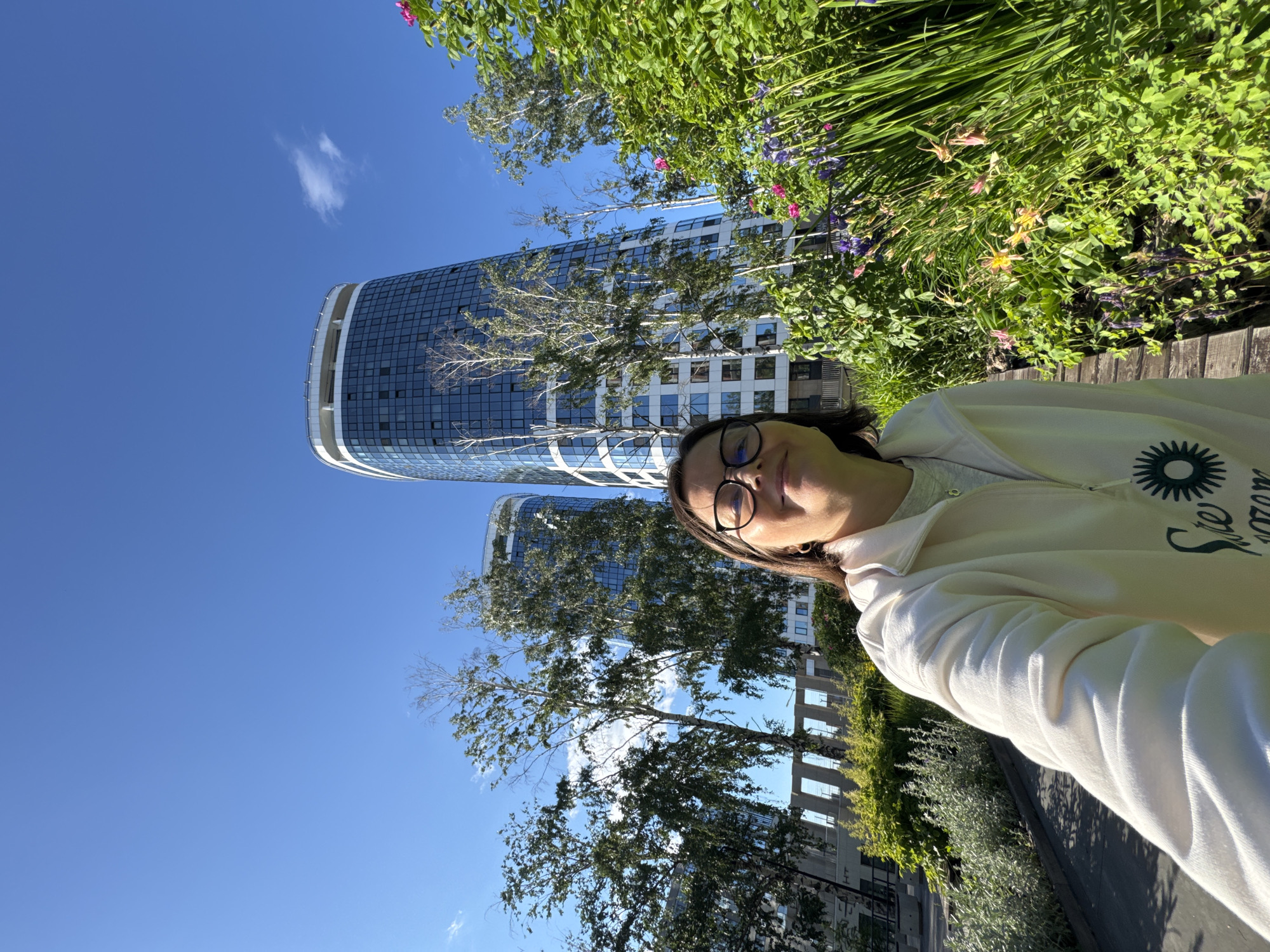 A person wearing a white hoodie and glasses takes a selfie outdoors on a sunny day. They are standing in front of a lush green garden with flowers and trees. In the background, two modern, curved glass skyscrapers rise against a clear blue sky. The architecture suggests a contemporary urban setting, possibly a park or residential area within the city. The specific location within Izhevsk, Russia, is not identifiable from the image alone, but the style of the buildings and the presence of greenery are characteristic of modern urban development.