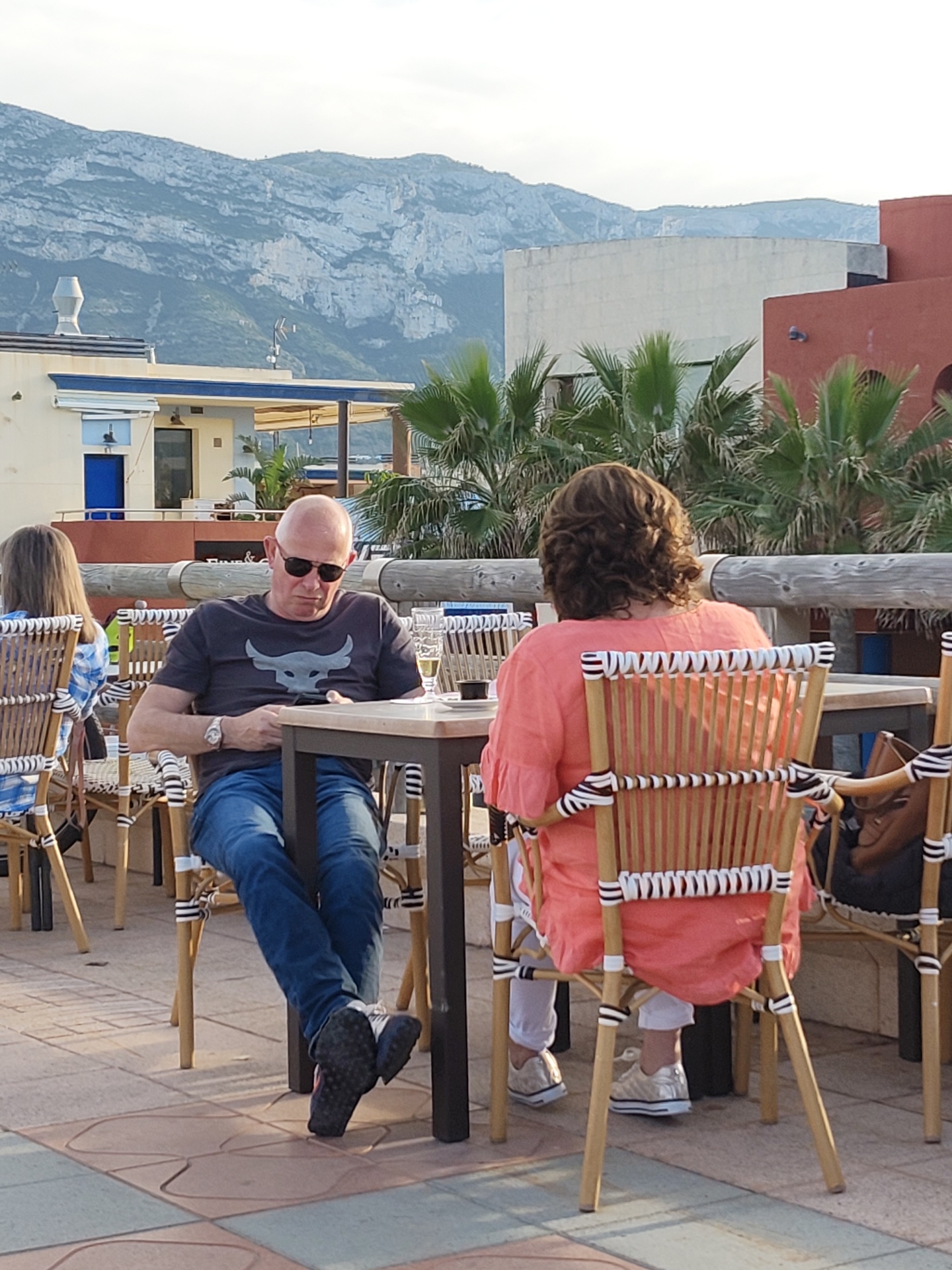 The image depicts an outdoor dining terrace in Dénia, Spain, with the prominent Montgó Massif visible in the distant background under a light sky.
In the foreground-middle ground, a bald male individual, wearing dark sunglasses, a dark grey t-shirt featuring a light grey bull's head logo, blue jeans, and dark athletic shoes with pink accents, is seated at a rectangular table. A wristwatch is visible on his left wrist. He is leaning slightly forward, directing his gaze downward at an unseen object in his hands. Opposite him, with her back to the camera, is a female individual with medium-length curly brown hair. She is wearing a coral-colored long-sleeved top, white trousers, and light-colored athletic shoes. A brown shoulder bag is suspended from the back of her chair.
On the table between these two individuals, a tall glass containing a light-colored liquid and a dark mug are visible. All visible chairs are of a woven cane or rattan design, featuring light-colored frames and black and white striped webbing on the backrests and seats. To the left, a portion of another female, wearing a blue and white checkered shirt, is visible seated at an adjacent table. The ground is paved with square tiles in varying shades of brown and blue.
A multi-layered wooden railing runs horizontally across the middle ground, separating the dining area from the background elements. Behind this railing, a light-colored building with blue trim, a blue door, and a chimney-like ventilation structure on its roof is visible. Further back, a stark white rectangular building stands adjacent to a terracotta-colored building featuring arched architectural details. Numerous palm trees are interspersed throughout the middle and background. The scene is illuminated by natural light, indicative of late afternoon or early evening.