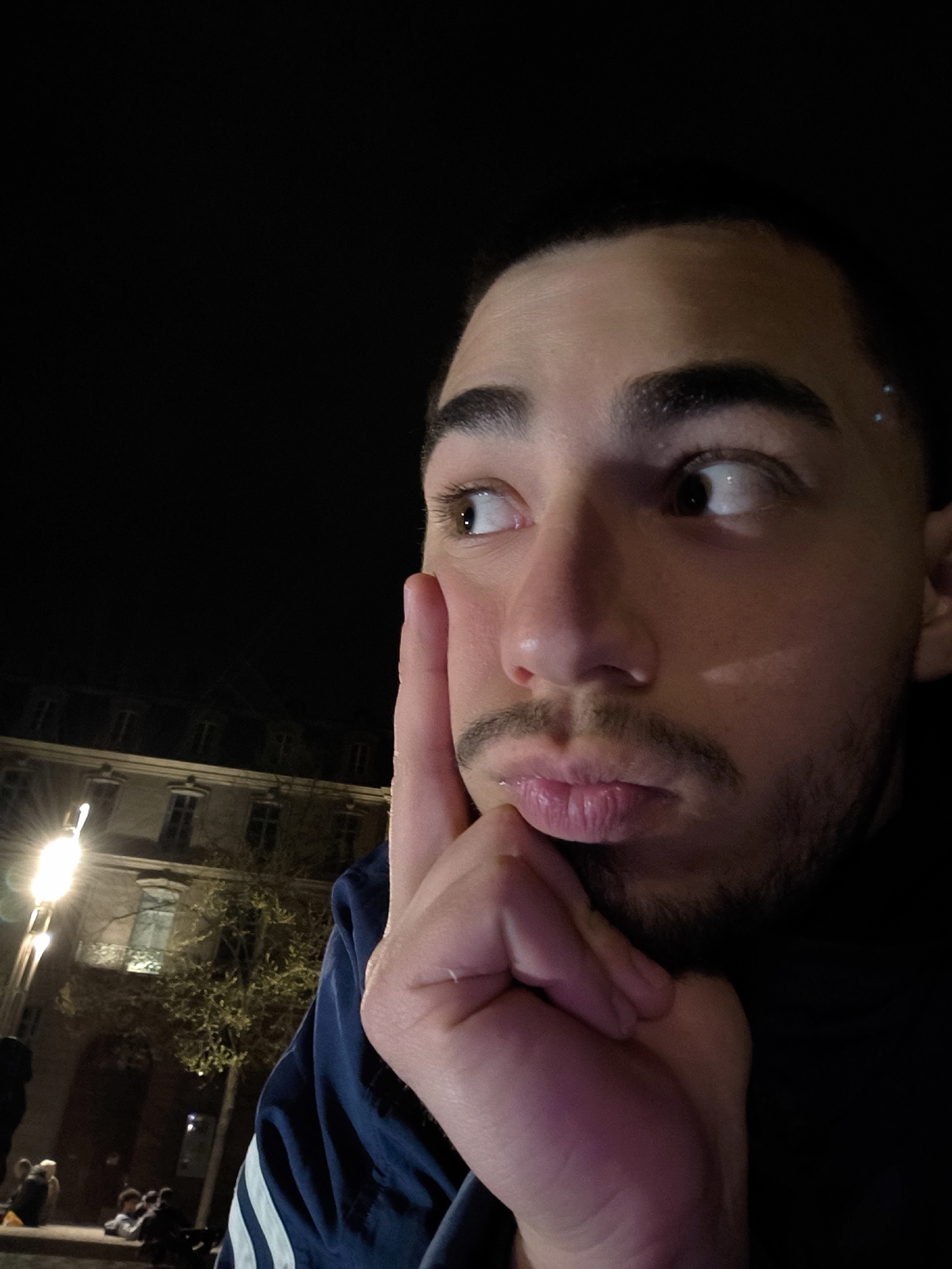 A selfie of a young man with dark hair, eyebrows, and a trimmed beard, looking slightly upwards and to the left, with his right index finger touching his cheek. He wears a dark blue jacket with three white stripes on the sleeve. In the blurred background, a building with multiple windows is illuminated by a street lamp. People are sitting on a bench in front of the building. This image was taken at night, in Strasbourg, France.