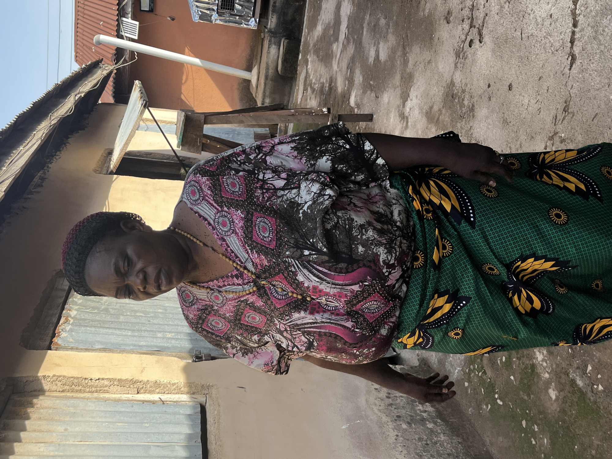A woman stands in an outdoor area. She wears a patterned top with pink, white, and black designs, a wooden beaded necklace, and a green and black patterned skirt. The background shows a building with corrugated metal walls, an orange building, and a concrete outdoor surface. The location appears to be Jos, Nigeria.