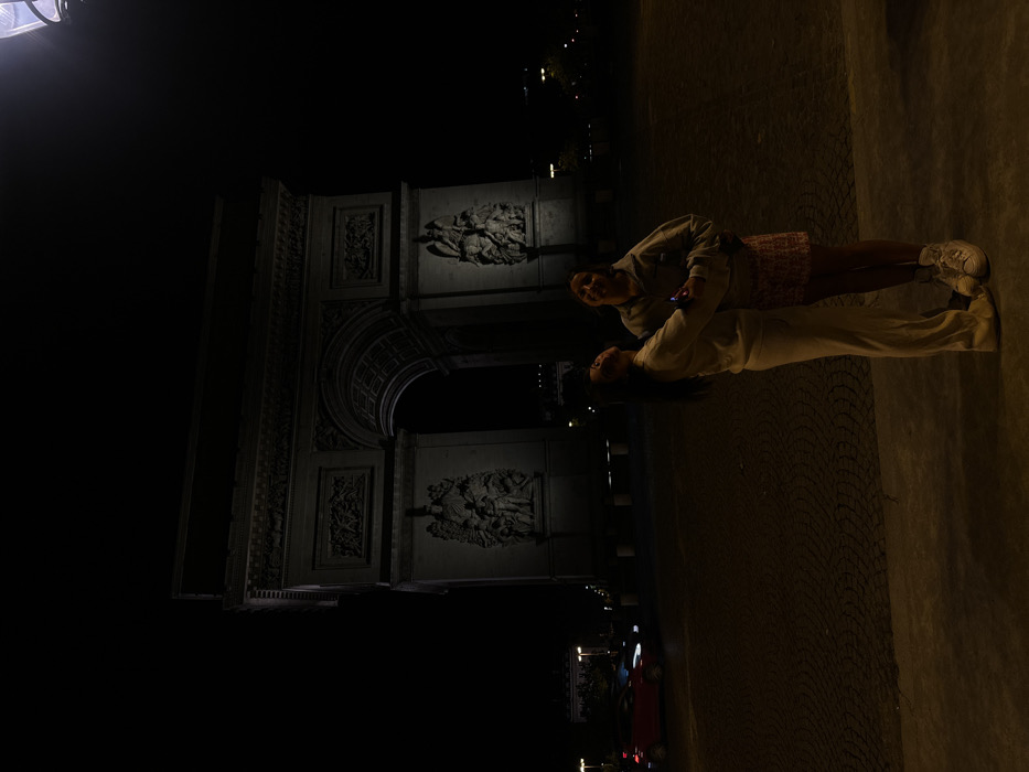 This nighttime image depicts two individuals positioned in front of the Arc de Triomphe in Paris, France, located at Place Charles de Gaulle. The monumental stone archway, adorned with classical architectural details and large relief sculptures, dominates the background, partially illuminated by artificial lighting.
In the foreground, two female subjects are closely positioned, appearing to embrace. The subject on the left, with long dark hair, wears a light-colored, possibly beige or off-white, matching tracksuit (sweatshirt and wide-leg trousers). She holds a smartphone in her left hand and looks upwards and slightly to her right with her mouth open. The subject on the right, shorter, wears a light-colored hooded sweatshirt, a patterned short skirt (red and white), and white chunky athletic shoes. She also looks upwards, with her mouth open and tongue extended in a playful manner. Their inner arms are wrapped around each other. The ground beneath them is dark and appears to be paved, possibly with cobblestones. In the far left mid-ground, a red vehicle with illuminated headlights is visible on a dimly lit street. The overall environment is dark, with the primary light sources emanating from the monument, a streetlamp at the upper right, and distant vehicle lights.