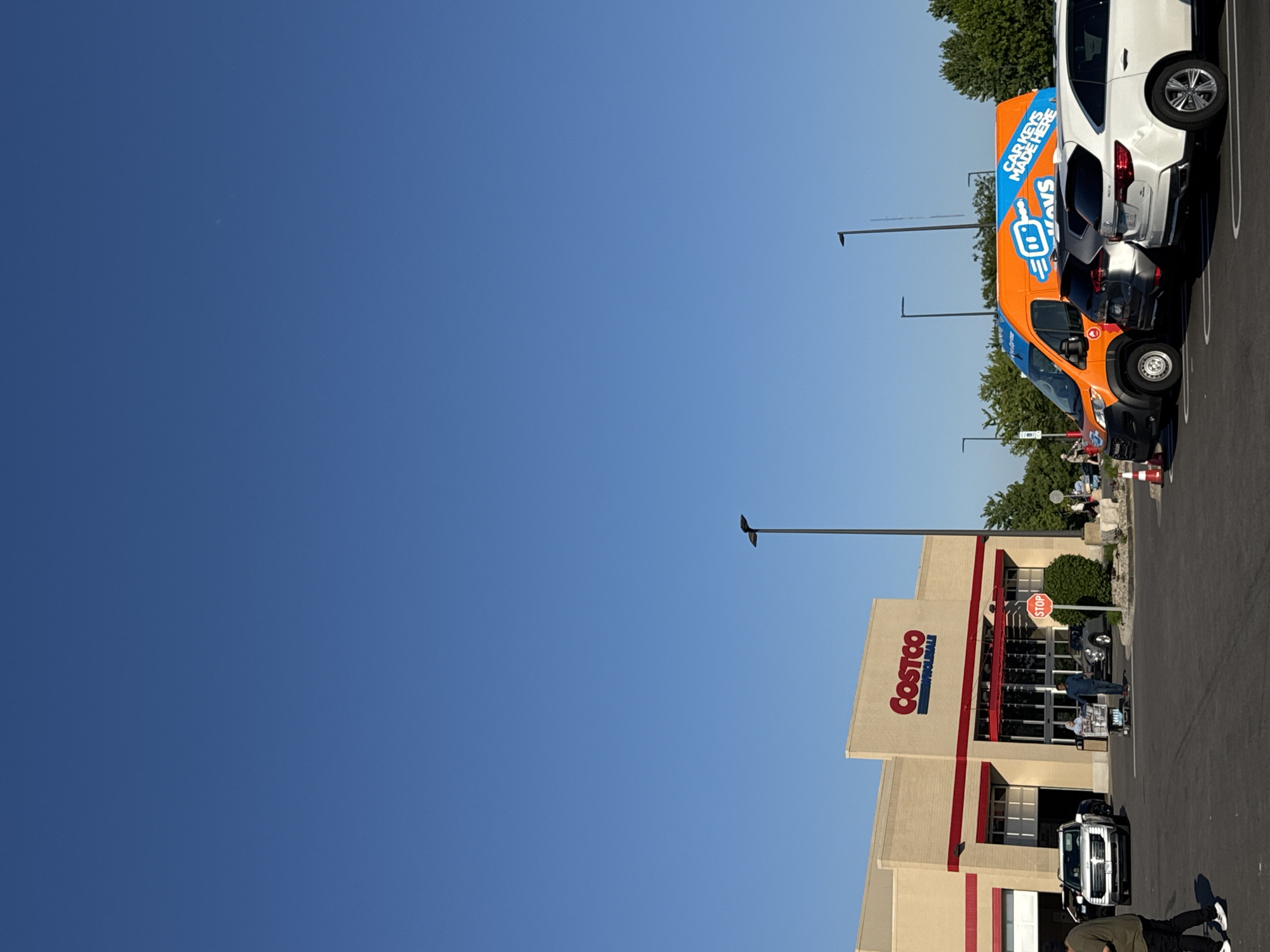 The image shows the Costco Wholesale store in Bloomingdale, Illinois, USA. The Costco building is beige with red trim around the entrance. Customers are visible near the entrance, some with shopping carts. In the parking lot, vehicles are parked, including an orange van with "Car Keys Made Here" printed on the side. The sky is clear and blue. Streetlights are visible. The image is taken during the day.
