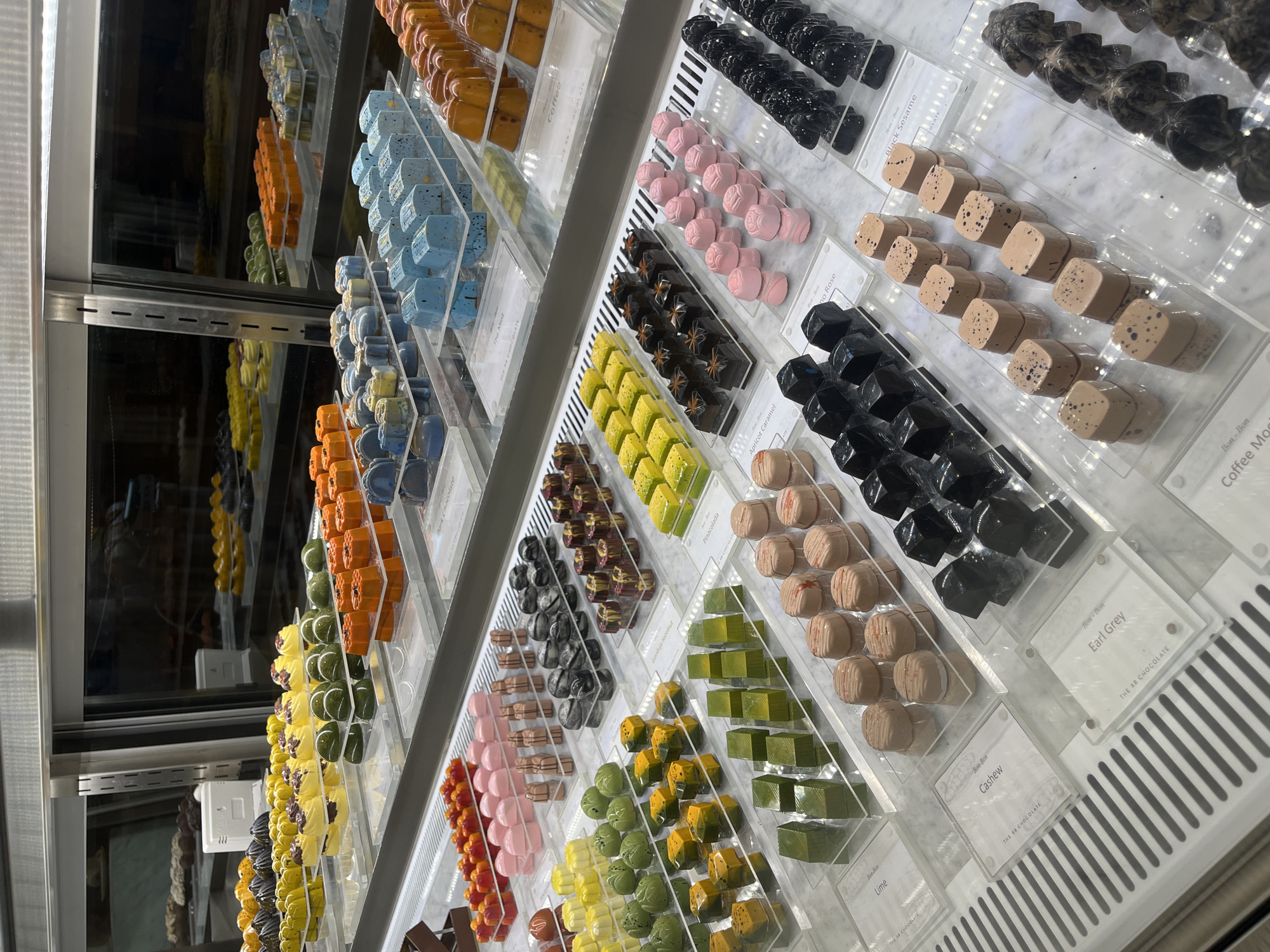 The image captures a vibrant and extensive display of gourmet bonbons arranged meticulously within a well-lit retail display case. The setting appears to be a high-end chocolate shop or confectionery in North Kuta, Indonesia. The chocolates are showcased on clear acrylic trays, organized in neat rows and columns across multiple shelves. A reflective back panel within the display case creates an illusion of greater depth and abundance, mirroring some of the colorful sweets. No people or activities are depicted, emphasizing the product itself. The time of day or external weather conditions are not visible, as the photo is taken indoors.
A wide array of bonbon flavors is suggested by the diverse colors, shapes, and textures. The chocolates come in various hues, including deep black, dark grey, pastel pink, beige, light blue, bright yellow, lime green, orange, and cream. Shapes range from perfect cubes and spheres to intricate rose-like designs and other molded forms. Many pieces are decorated with speckles, swirls, or textured patterns.
Visible text on small white labels indicates specific flavors and the brand name. Identified flavors include "Coffee," "Pinacolada," "Apricot Caramel," "Rose," "Black Sesame," "Lime," "Cashew," "Earl Grey," "The Mint," and "Dark Chocolate." The brand "THE RR CHOCOLATE" is consistently featured on the labels, often accompanied by the term "Bon Bon," denoting the type of confectionery. The display creates an appealing and sophisticated presentation of artisanal chocolates.