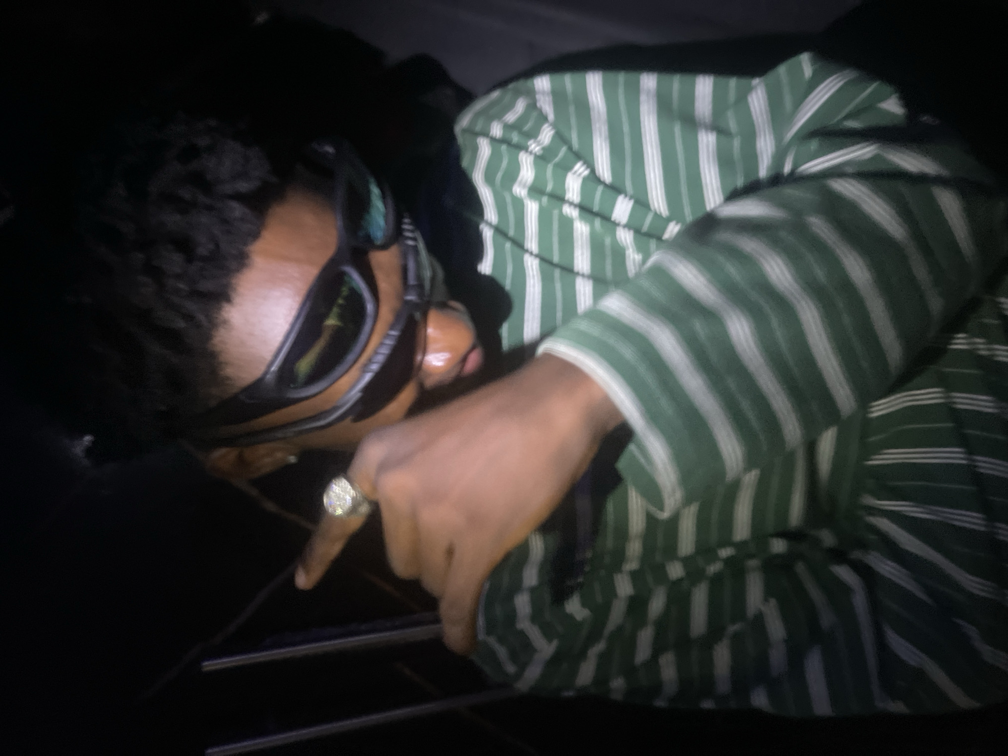 A person wearing sunglasses and a green and white striped shirt is pointing down with their right index finger. They are also wearing a large ring. The background is dark. There is no verifiable evidence to place the location of the photo within Lokoja, Nigeria.