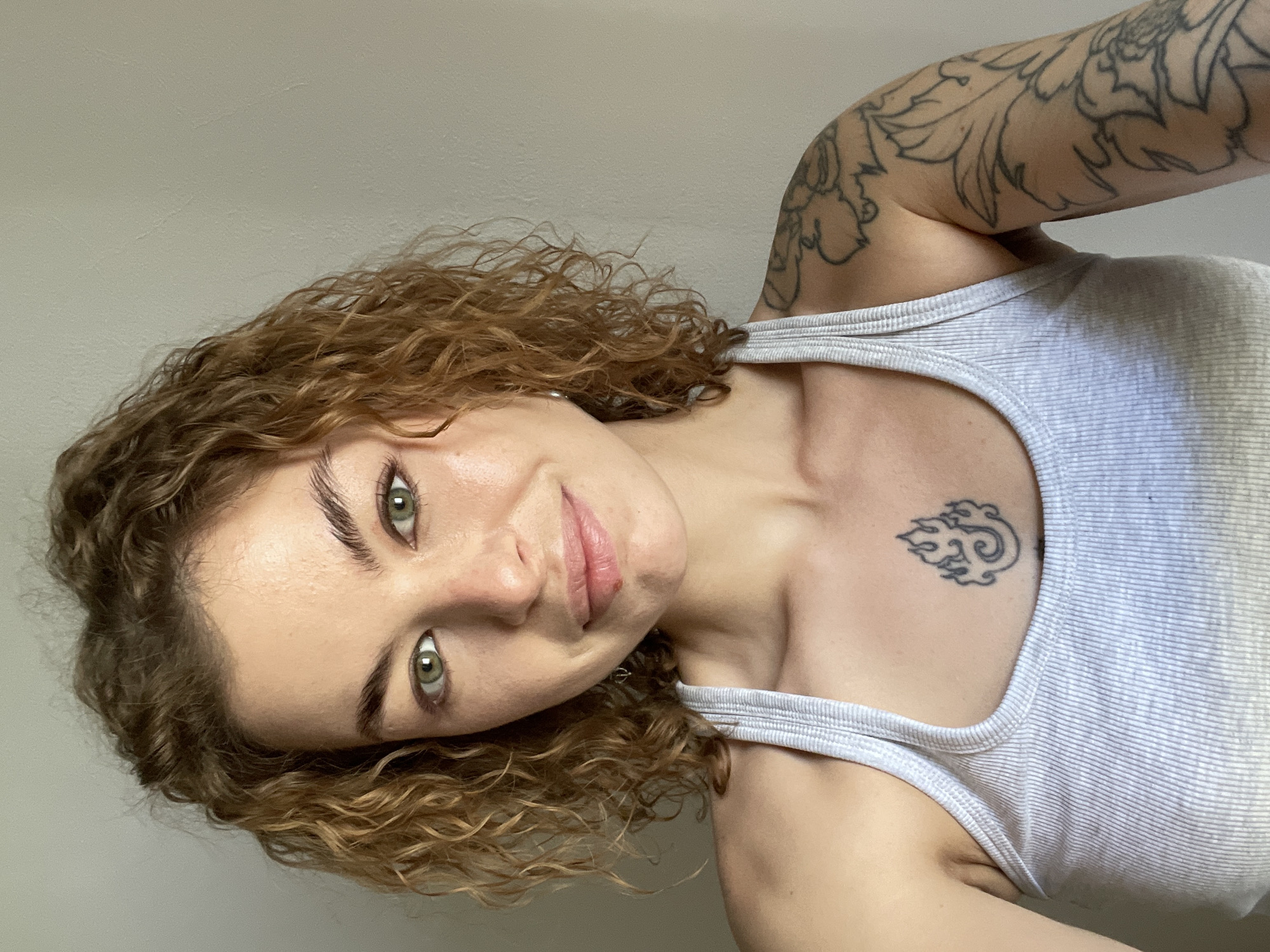 A woman is angled, facing the camera. She has curly, light brown hair and green eyes. She is wearing a light gray tank top. She has tattoos on her upper arm and chest. The background is a plain, light-colored wall. The location is not discernible from the image.