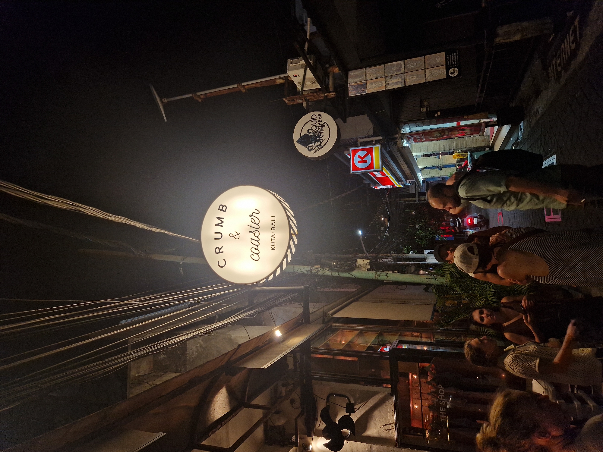 The image depicts a nighttime street scene in Kuta, Bali, Indonesia. The signage for "CRUMB & COASTER, KUTA-BALI" is prominently displayed, illuminated as a circular sign above a building front with visible framing and a black ceiling fan. Wires are strung across the upper portion of the frame. A smaller sign for "SQUID," also in Kuta, Bali, hangs nearby, along with other business signs, including an Indomaret store. Several people are present on the street, some appear to be interacting with each other in a casual manner. One individual wears a backpack and stands near the edge of the street. The background shows dim streetlights and vehicle headlights indicating street traffic.