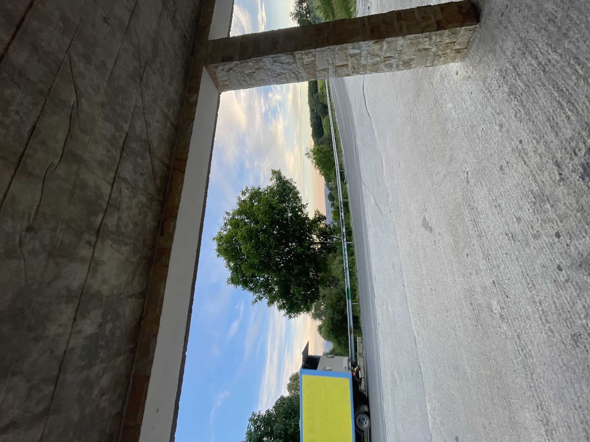 A wide-angle, eye-level outdoor shot captures a serene landscape with a focus on a road and a truck, framed by a concrete overhang and a stone pillar on the right. The scene appears to be located on the outskirts of Valkosel, Bulgaria.
In the foreground, a rough, concrete surface extends towards the viewer, showing some texture and scattered dark spots. To the right, a thick, square pillar constructed from light-colored, rough-hewn stones stands prominently, supporting the overhead structure. The overhang above is made of gray concrete, with visible lines and imperfections.
Beyond the foreground, a paved road curves gently through a verdant landscape. On the left side of the road, a truck with a bright yellow cargo box and a blue trim is visible, moving away from the viewer and towards the right. The truck is partially obscured by its motion blur.
The middle ground features a cluster of lush green trees, with one particularly large, mature tree with a full canopy dominating the center of the frame. A silver guardrail runs along the side of the road, separating it from the rolling hills in the background.
The sky above is a picturesque blend of soft blue with scattered white and wispy clouds, tinged with the warm hues of either sunrise or sunset. The overall atmosphere is tranquil and natural, suggesting a rural or semi-rural setting. The presence of the truck and the unfinished concrete structure hint at ongoing development or activity in the area. There are no people visible in the image.