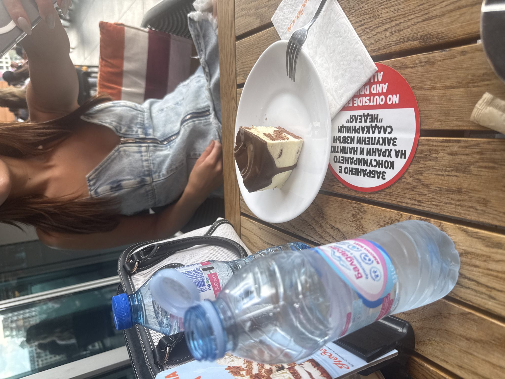 The image captures a casual cafe setting, seen from an overhead perspective, rotated 90 degrees clockwise. A person, partially visible on the left, wears a light blue denim top and has long brown hair. Their arm and part of their torso are visible.
In the foreground and center, a wooden table holds various items. A white plate features a slice of layered cake, possibly a tiramisu or similar dessert, topped with chocolate sauce and sprinkles. A fork rests on a white napkin beside the plate. Next to these, a striking red circular sign is prominently displayed. Below the table level, what appears to be a black and white handbag is partially visible.
Two plastic water bottles are present, one larger and clearer in the foreground, labeled "Балдара" (Baldara) and "500mle," and another smaller bottle slightly behind it. A menu or coaster below the water bottles also shows the word "Неделя" (Nedelya). In the background, beyond the person, a cushion with orange and white stripes is visible, suggesting cafe seating. Reflections in a window or glass partition hint at a cityscape outside.
The red sign, primarily in Cyrillic, translates to "CONSUMPTION IS FORBIDDEN FOR FOOD AND DRINKS PURCHASED OUTSIDE OF "NEDELYA" PATISSERIES." A partial English translation, "NO OUTSIDE FOOD AND DRINKS ALLOWED," is also visible. This text, combined with the "Nedelya" branding and the "Baldara" water bottle (a Bulgarian brand), confirms the location as Sofia, Bulgaria, likely at a Nedelya Patisserie, a well-known chain in the country. The scene depicts a daytime outing, possibly for a dessert break.