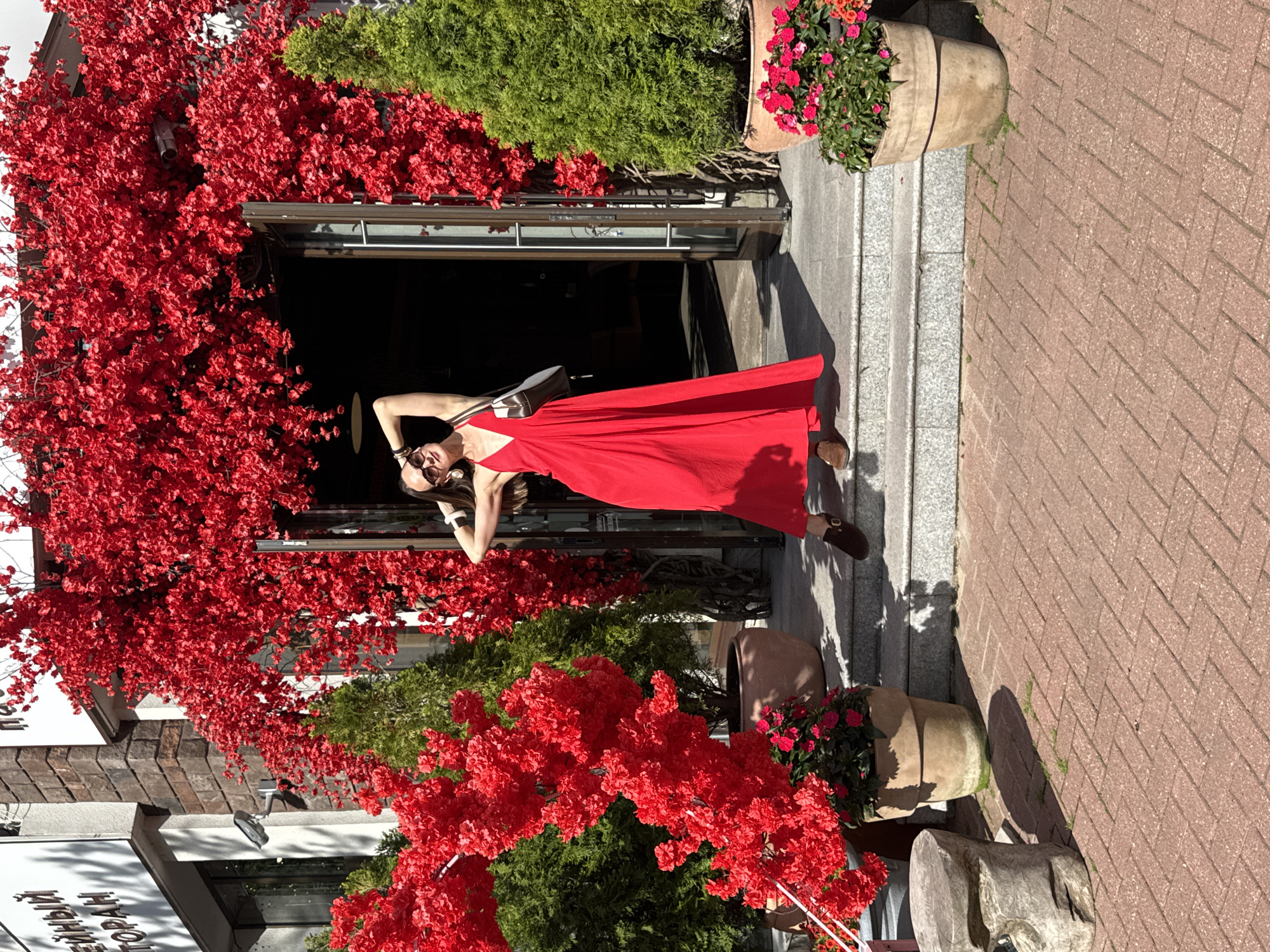 A woman in a red dress poses in front of a building adorned with red floral decorations. The woman is standing on a set of stone steps. She is wearing sunglasses and has her right arm raised behind her head. A handbag is slung over her left shoulder. To the left and right of the entrance, there are green conical trees in pots. The ground in front of the steps is paved with red brick. The building appears to be a restaurant or cafe, with signage visible above the entrance. The overall scene suggests a warm, sunny day in an urban or semi-urban setting.