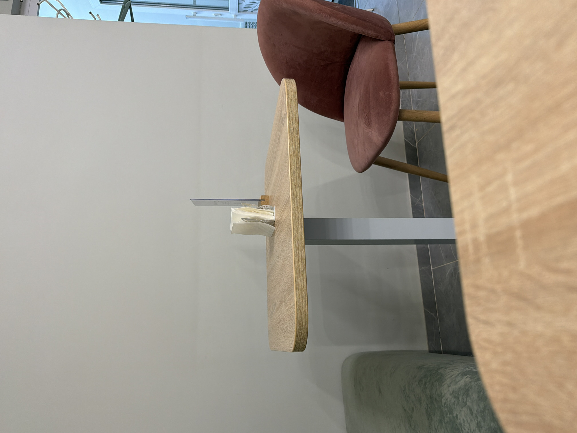 The image shows a close-up view of a small cafe or restaurant setting. In the foreground, a light-colored wooden table is partially visible. Beyond this table, a square wooden table with rounded edges is the central focus. On this table, a holder with white napkins stands. To the right of the table, a plush, rust-colored chair with wooden legs is positioned. To the left of the table, a light green upholstered bench is visible. The background consists of a plain, light grey wall. The flooring is dark grey tile with a subtle marbling pattern. The overall impression is a clean and minimalist interior. The image does not contain any people or explicit actions. The scene is likely located in the city of Фролы, Russia, based on the prompt.