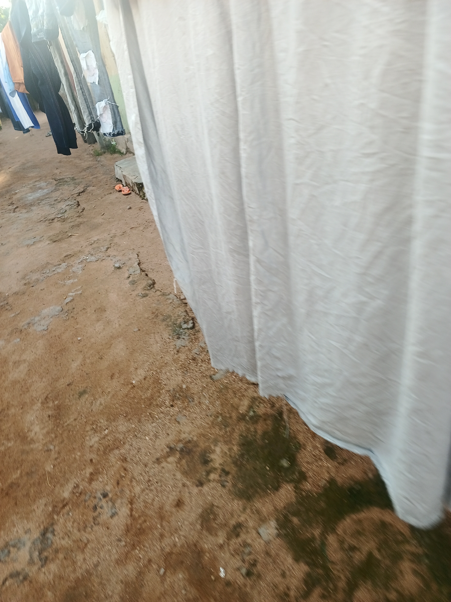 This media file depicts an outdoor scene in Jalingo, Nigeria, likely during the daytime given the bright lighting. The primary focus is on a clothesline strung with various garments, including what appear to be dark-colored pants, a gray pair of ripped jeans with white patches, and lighter-colored items like a blue and white garment. A large, white piece of fabric, possibly a sheet or curtain, is draped prominently in the foreground, obscuring some of the other items on the line and dominating the right side of the frame.
The ground beneath the clothesline is a mix of dry, reddish-brown earth with patches of lighter colored soil and some darker, possibly damp or mossy areas. There are some small stones and debris scattered on the ground. In the middle ground, towards the left side of the image, a pair of bright orange flip-flops are visible on the ground, suggesting a casual, lived-in environment. The overall impression is of a domestic setting where laundry is being air-dried. There are no people visible in the frame, and no text is present. The weather appears to be clear and sunny, facilitating the drying of clothes.