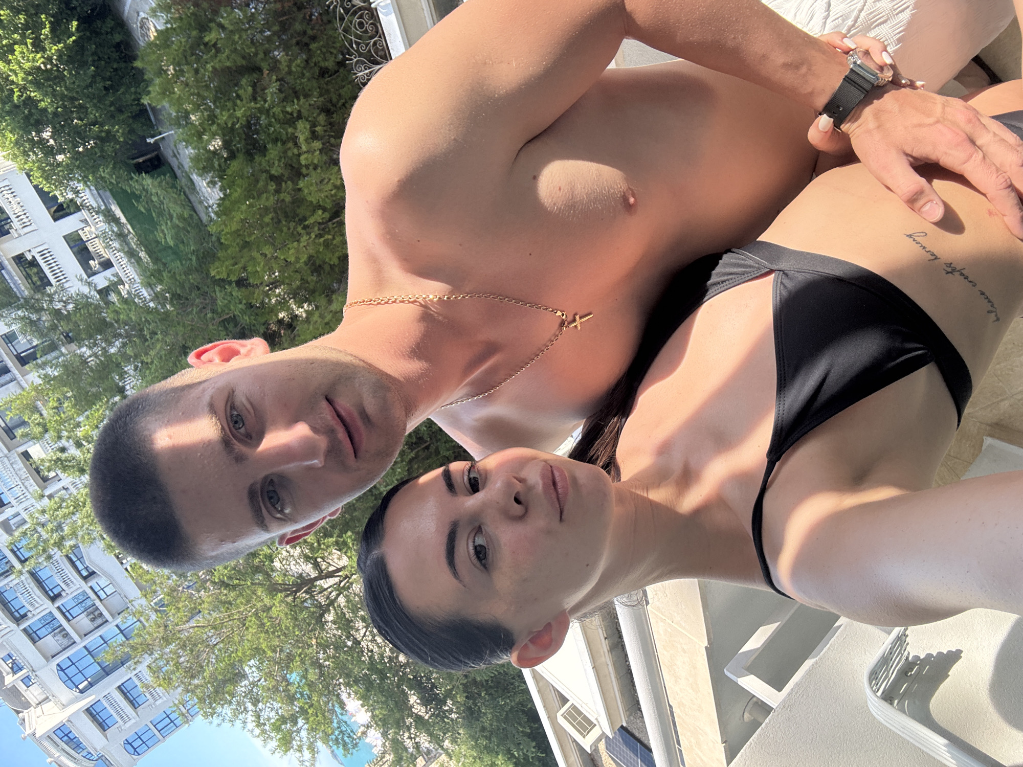 The image shows a close-up of two people, a man and a woman, in what appears to be a sunny outdoor setting. The man is in the foreground, looking towards the camera with a slight smile. He is shirtless and wearing a gold chain with a cross pendant. The woman is positioned behind and to the side of the man, her face turned towards him. She is wearing a black bikini top. Her arm is resting on the man's chest, and she has a tattoo on her side. A watch is visible on the man's wrist. In the background, there are green trees and a building with multiple balconies, suggesting an urban or resort location. The lighting indicates it is daytime, likely a sunny day given the shadows and bright illumination on the subjects. The overall mood appears relaxed and intimate. There is no visible text in the image.