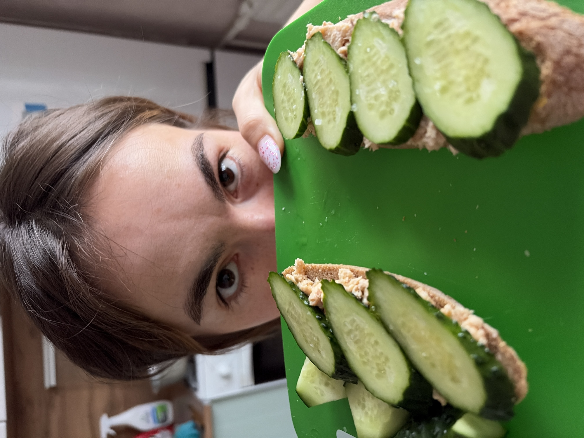 A photograph shows a woman holding a green cutting board with two open-faced sandwiches topped with cucumber slices. The woman's face is visible in the left part of the frame, with her eyes looking directly at the camera. She has brown hair and eyebrows, and her fingernails have a pink and white design. The sandwiches consist of a spread (possibly tuna or another fish mixture) on bread, with several cucumber slices arranged on top. The background shows a glimpse of what appears to be an interior space with shelves, containers, and other household items. The location is not evident from the image.