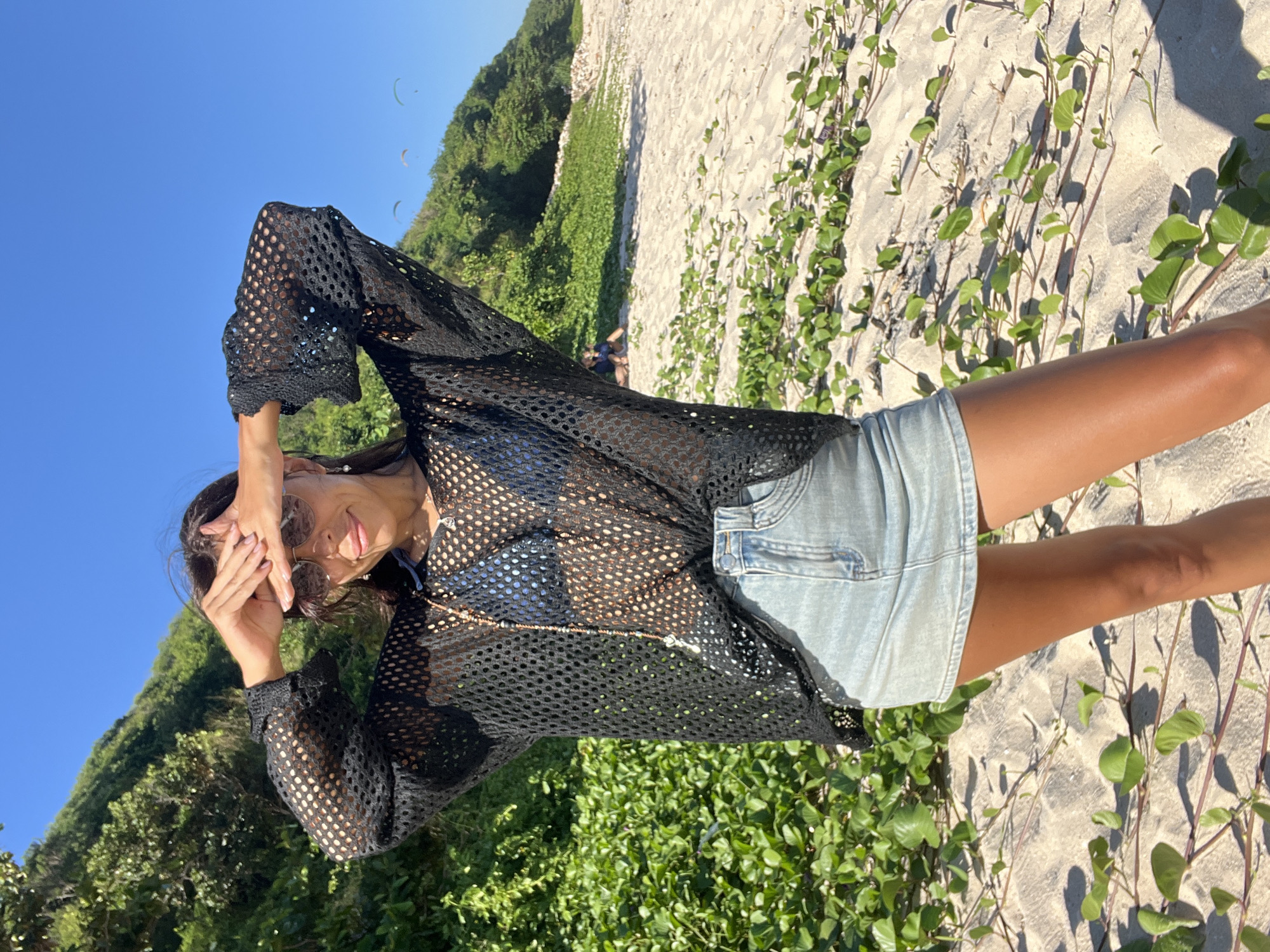 A young woman stands on a sunny beach in Kuta Selatan, Indonesia, posing playfully for the camera. She is seen wearing a black mesh long-sleeve top, a light blue denim skirt, and sunglasses. Her dark hair flows around her shoulders as she holds her hands to her forehead, shielding her eyes from the bright sun, with a slight smile and her tongue playfully sticking out.
The setting is a sandy beach with patches of creeping green vegetation. In the background, a lush green hill or cliff rises prominently. Further back, the clear, vibrant blue sky is visible, dotted with three paragliders soaring in the distance. Another individual is faintly visible, seated or crouching on the sand near the base of the hill. The bright, direct sunlight and absence of clouds indicate a clear and pleasant day, likely late morning or early afternoon. The scene captures a relaxed moment of leisure on a tropical coast, with recreational activities like paragliding taking place.