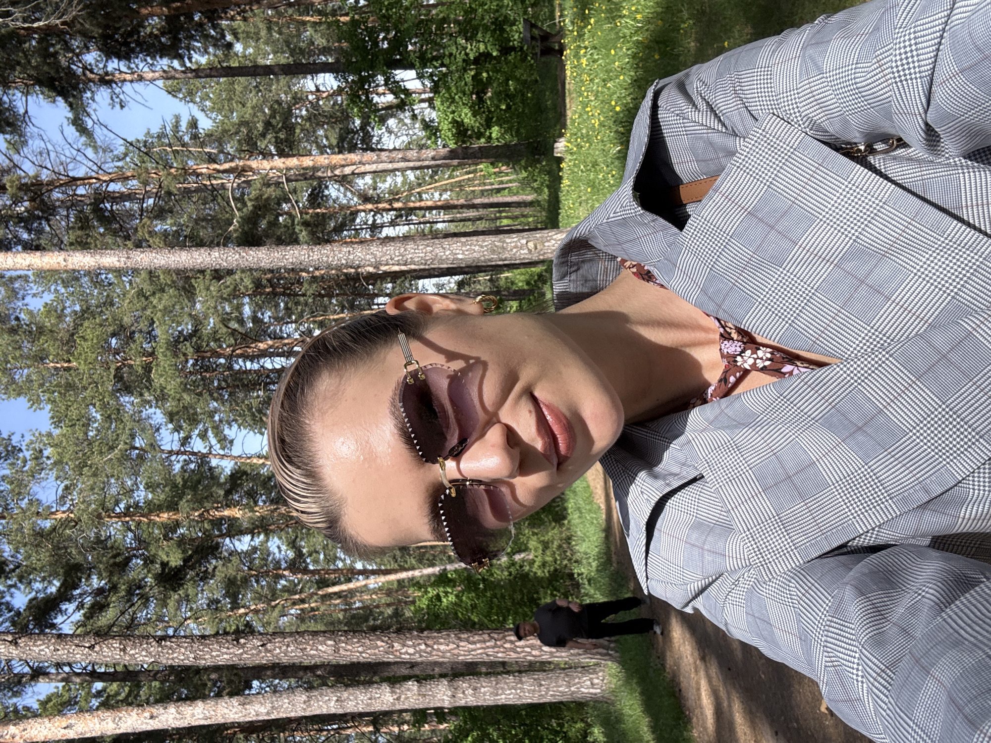 A selfie taken outdoors shows a person wearing rimless sunglasses with a brown tint and a grey plaid blazer. A glimpse of a floral-patterned top can be seen underneath the blazer. The person has fair skin and their hair is slicked back. Behind the person, a grove of tall pine trees is visible, with a clear blue sky peeking through the branches. On the ground, a path extends into the background, where a figure dressed in black is walking away. The location appears to be in a wooded area.