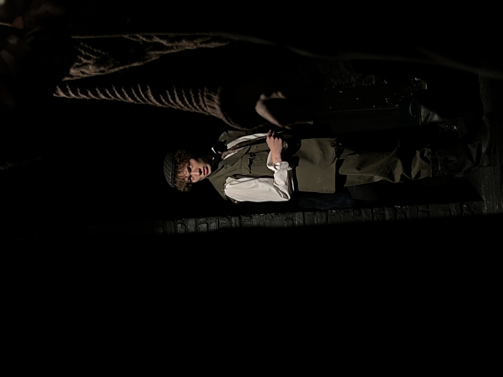 A person wearing a cap, white shirt, and vest lies on what appears to be an elevated surface made of wood or stone. A thick, dark rope hangs overhead. The scene is dimly lit, and the location is unknown.